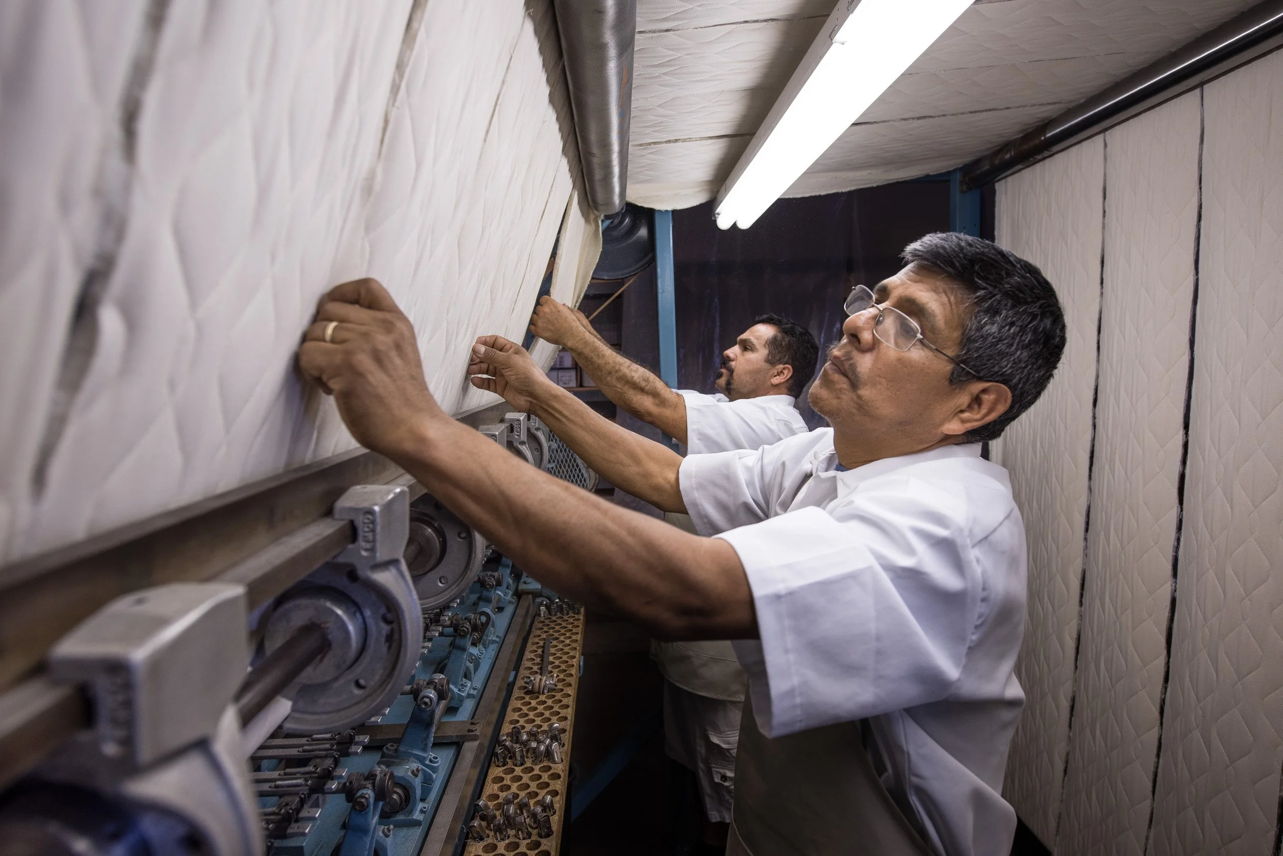 Two men in white lab coats working with machinery on a blue industrial machine in a room with quilted wall pads.
