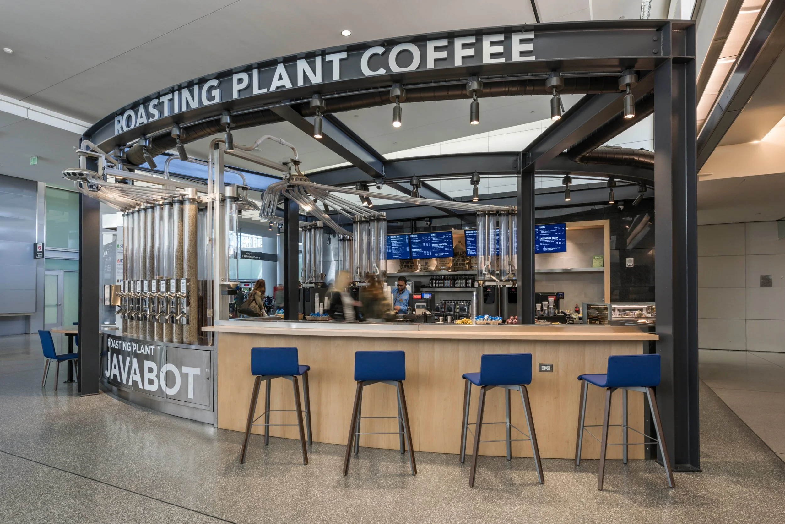 Coffee kiosk labeled 'Roasting Plant Coffee' inside a modern airport terminal with people and seating area.