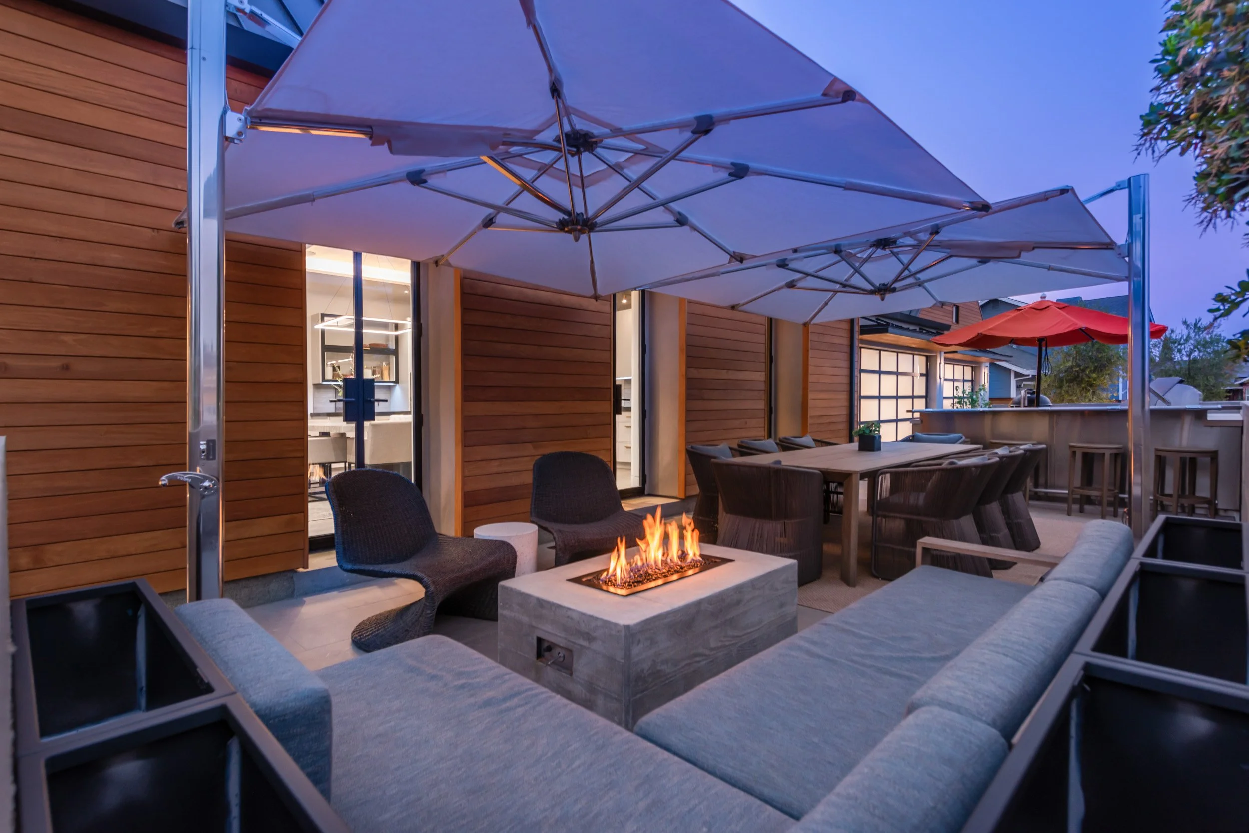 Outdoor patio area with a fire pit, seating, large umbrellas, and a wooden house in the background during dusk.