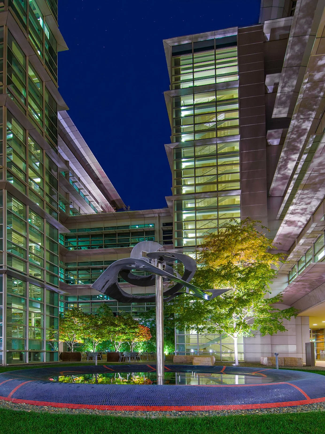 Night view of modern glass office buildings with illuminated interior, a courtyard with trees and benches, a decorative sculpture, and a circular water feature in the foreground.