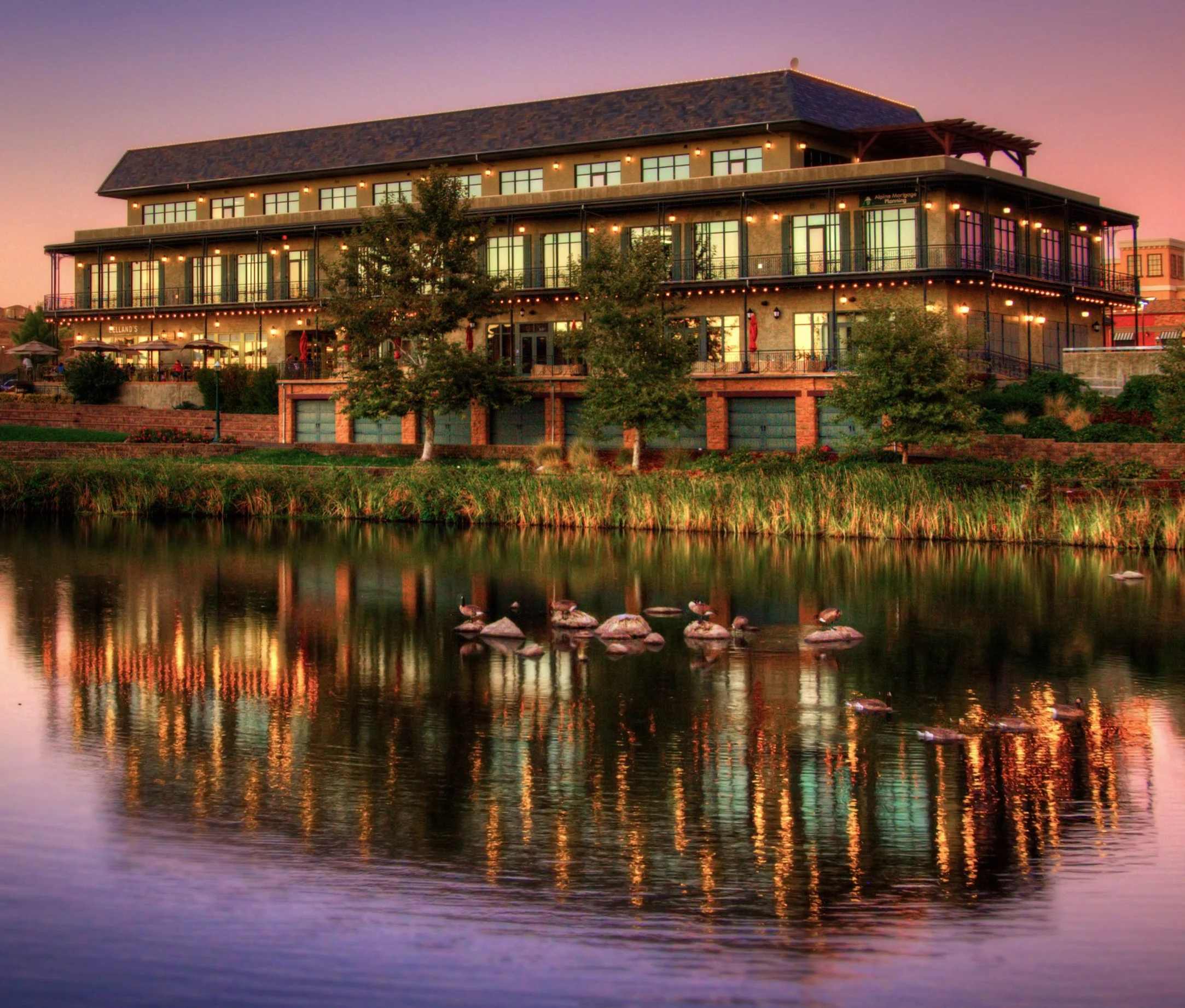 A multi-story building illuminated at dusk, featuring large windows, balconies, and warm exterior lighting, situated beside a calm body of water with ducks and trees in the foreground.