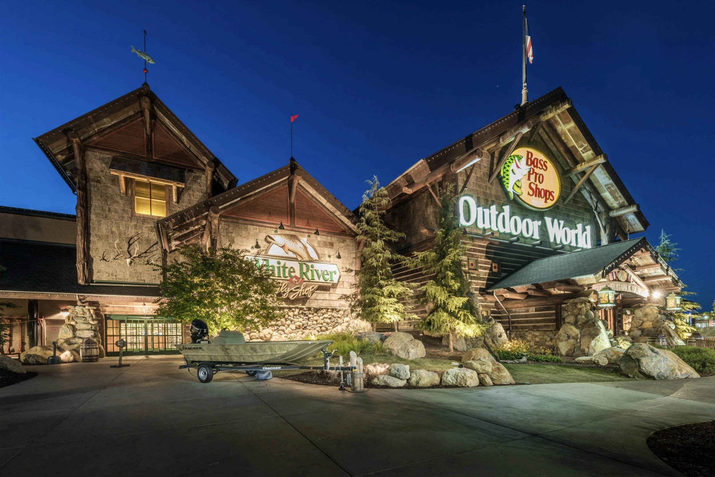 The exterior of a Bass Pro Shops outdoor world store at night, with illuminated signs, trees, rocks, a boat, and a clear evening sky.