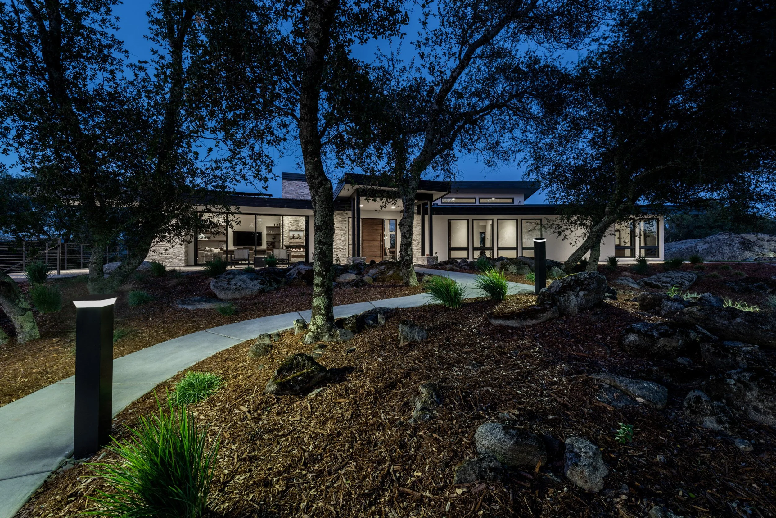 Modern house at night with large windows, surrounded by trees and landscaped garden with pathway and outdoor lighting.