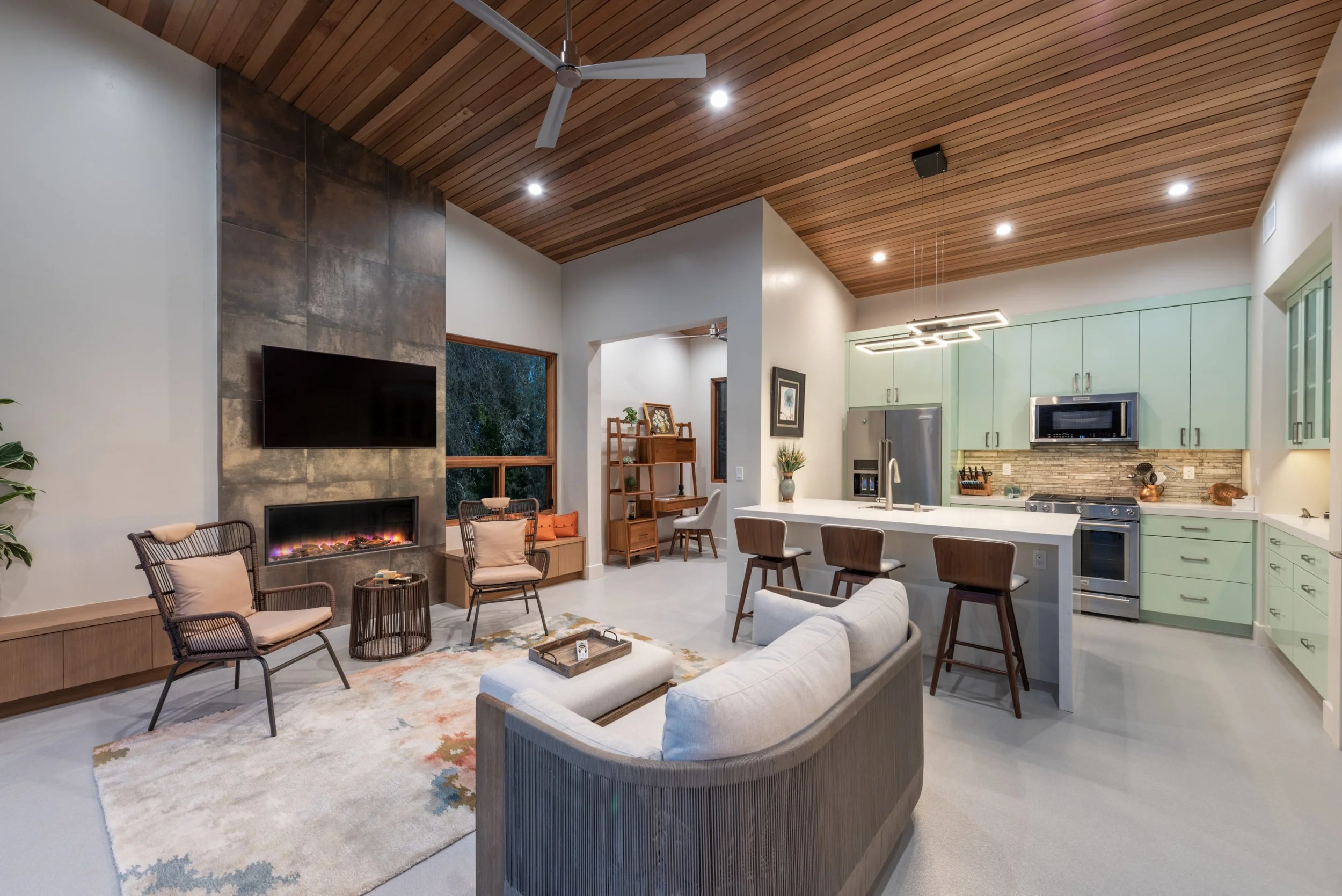 Interior view of a modern living room and kitchen with a wooden ceiling, fireplace, wall-mounted TV, dining area, and pastel green kitchen cabinets.