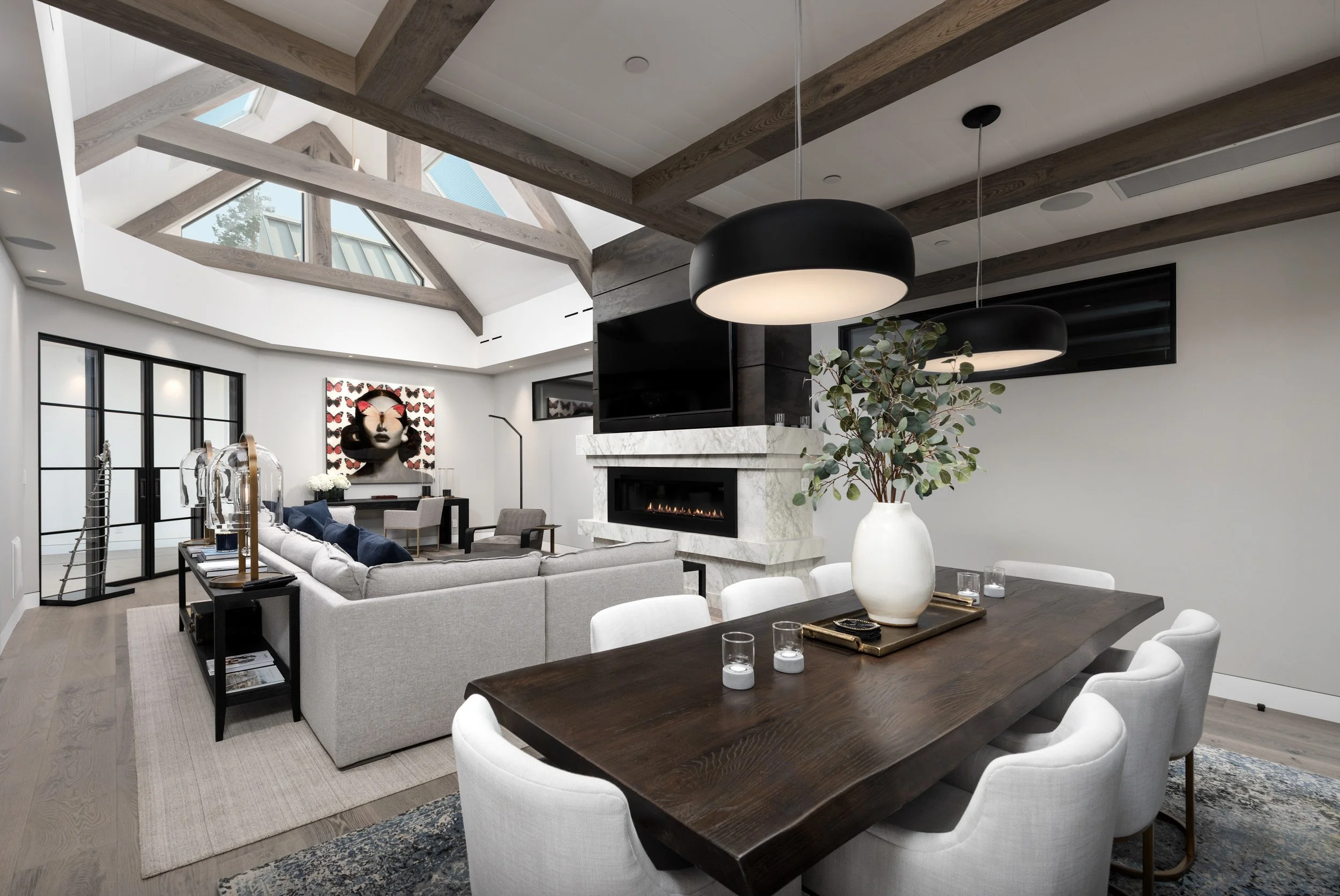 Modern living room with a beamed ceiling, skylights, and large windows, featuring a fireplace, artwork, and contemporary furniture.