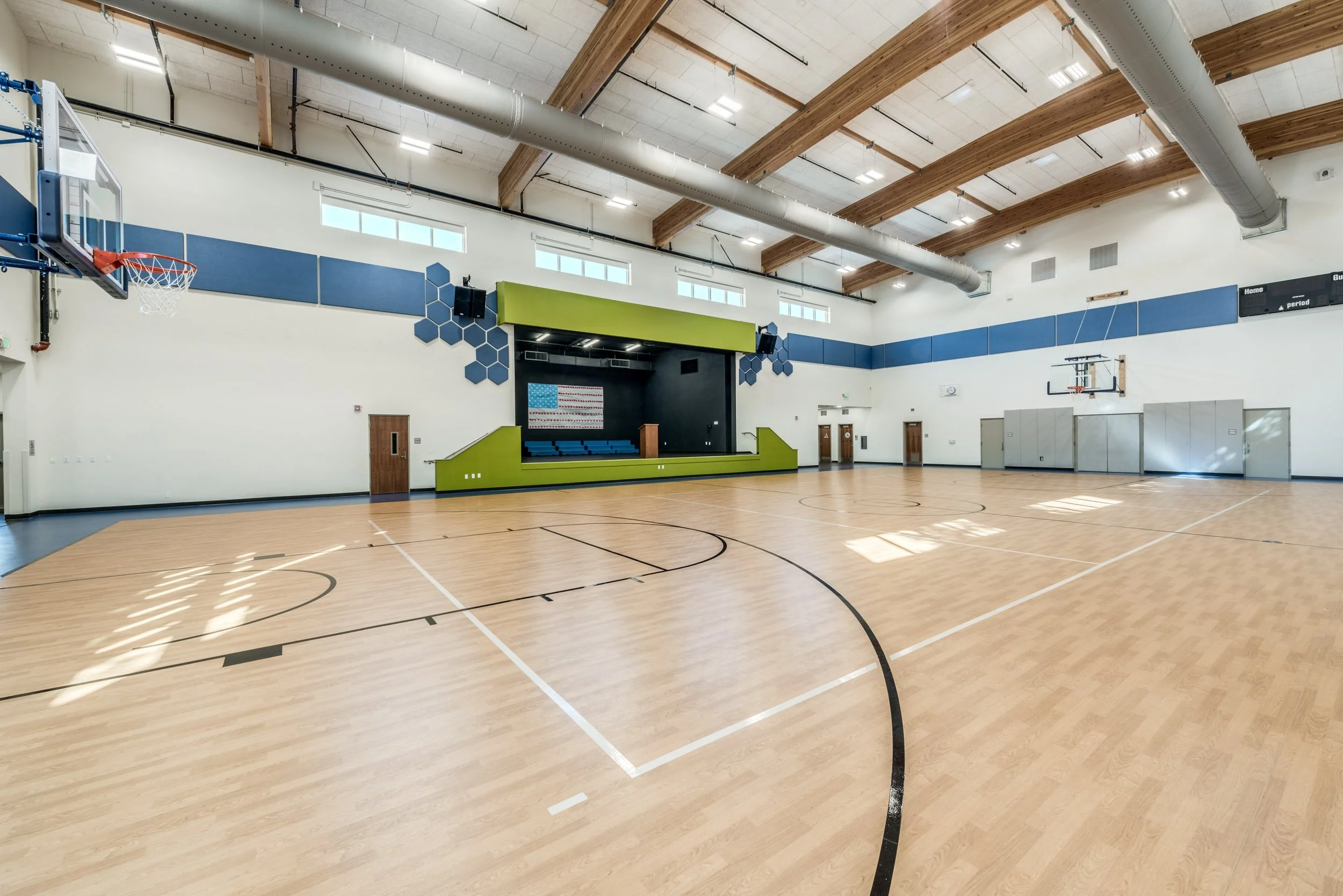 Empty indoor basketball court with wooden flooring, basketball hoops, and a stage with an American flag in the background.