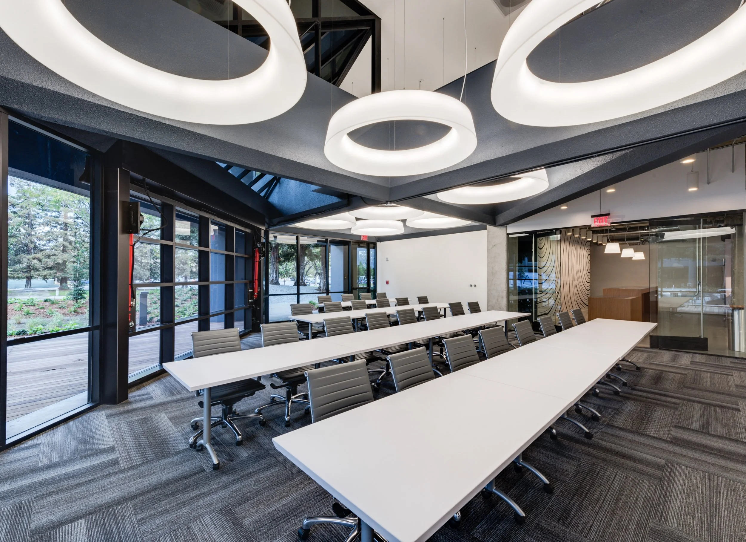 A modern conference room with large windows showing trees outside, white tables arranged in a U-shape, and gray chairs. Overhead lighting consists of circular pendant lights, and the room has a contemporary design with glass walls and a decorative wo