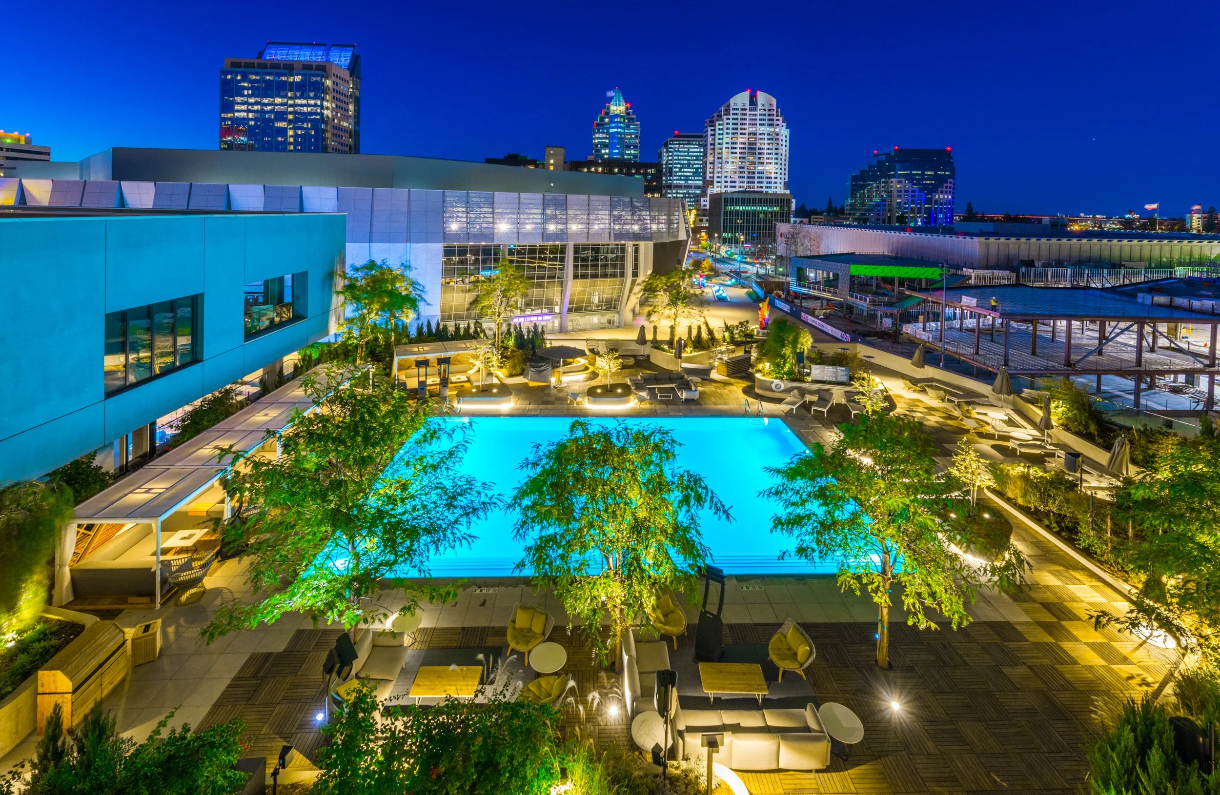 Night view of a cityscape showing tall buildings with illuminated windows, a pool area with lounge chairs and tables, and surrounding greenery, all brightly lit against a dark blue sky.
