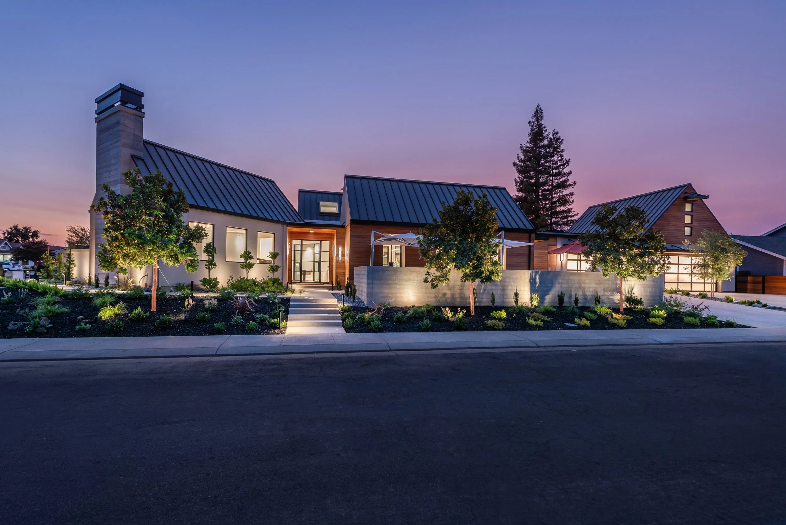 Modern house with a blue metal roof, large windows, and a well-maintained front yard with small trees and plants. The house is illuminated from inside, and the sky is at dusk with a gradient from purple to blue.