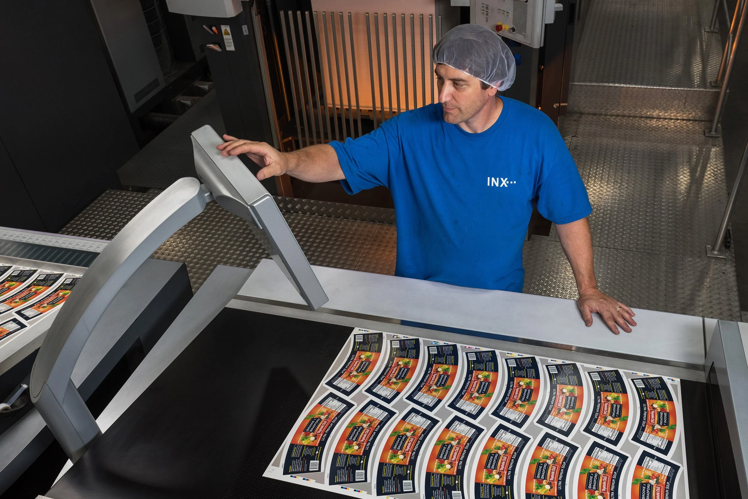 A man wearing a blue shirt and a hairnet operates a printer printing multiple copies of a colorful flyer or brochure. The printing machine is large and metallic, and the man is reaching out to adjust or interact with the machine.