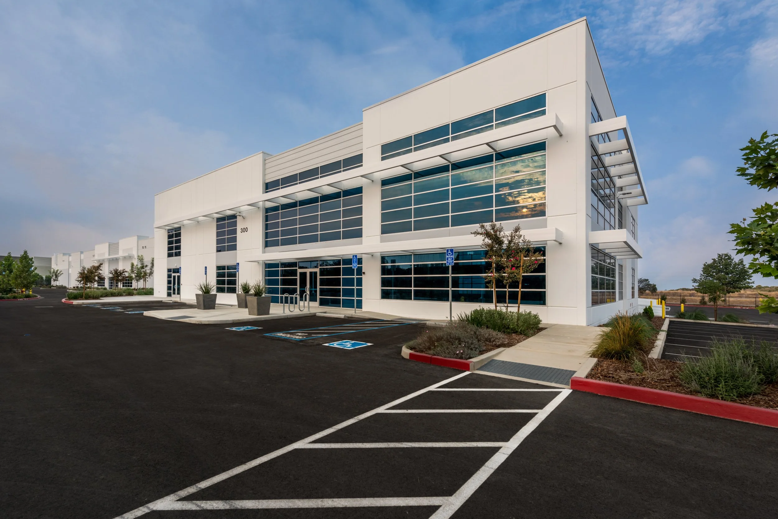 Modern white office building with large glass windows and an empty parking lot in the foreground. The sky is partly cloudy, and there are small trees and landscaping around the building.