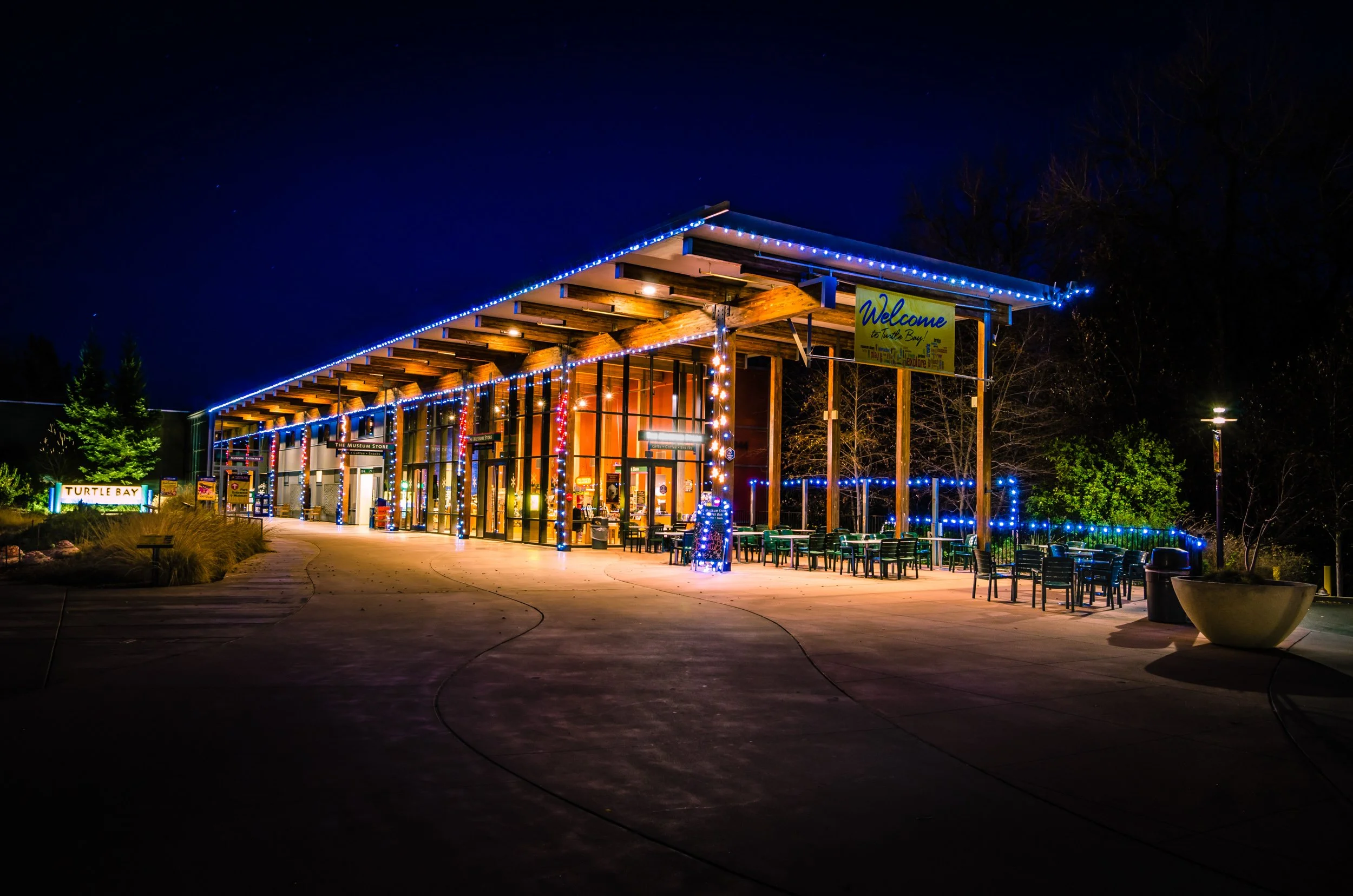 Night view of a building with a sloped roof decorated with blue string lights, outside seating area with chairs, trees, and illuminated signs, including a yellow welcome sign for Turtle Bay.