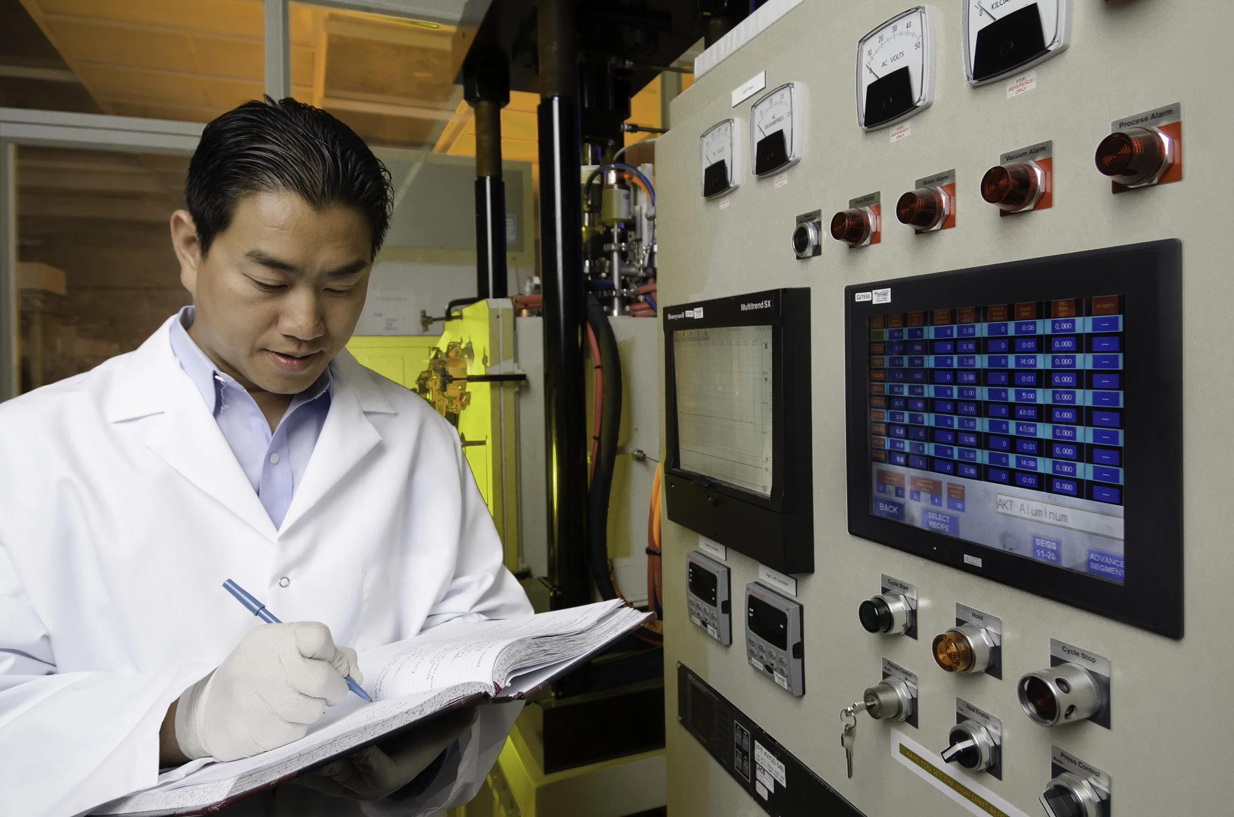 A scientist in a white lab coat taking notes while working on electrical equipment with gauges and digital screens in a laboratory.