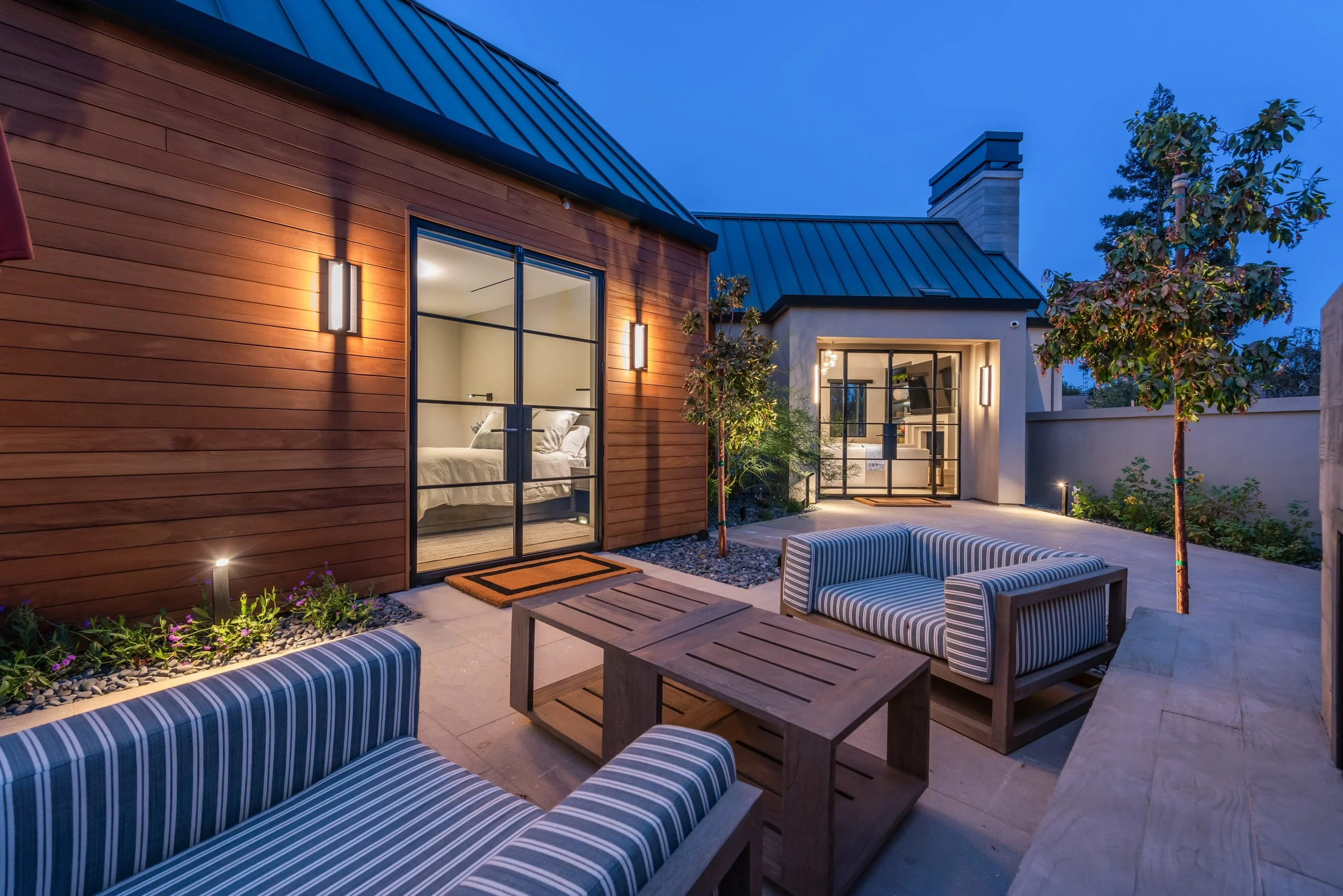 Modern house exterior at dusk with outdoor seating area, striped cushions, small trees, and illuminated windows.