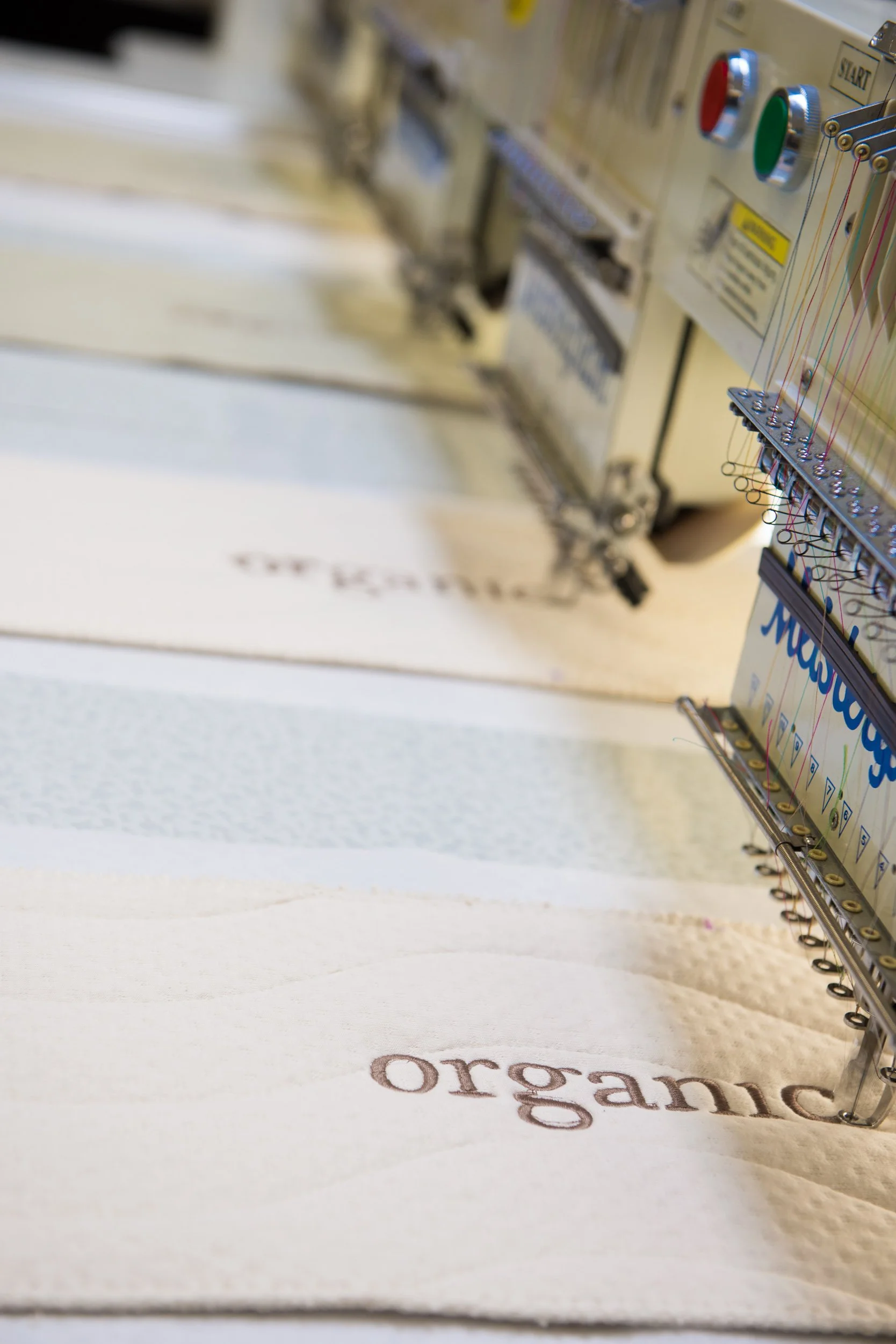 Close-up of embroidery machine stitching the word 'organic' onto beige fabric.