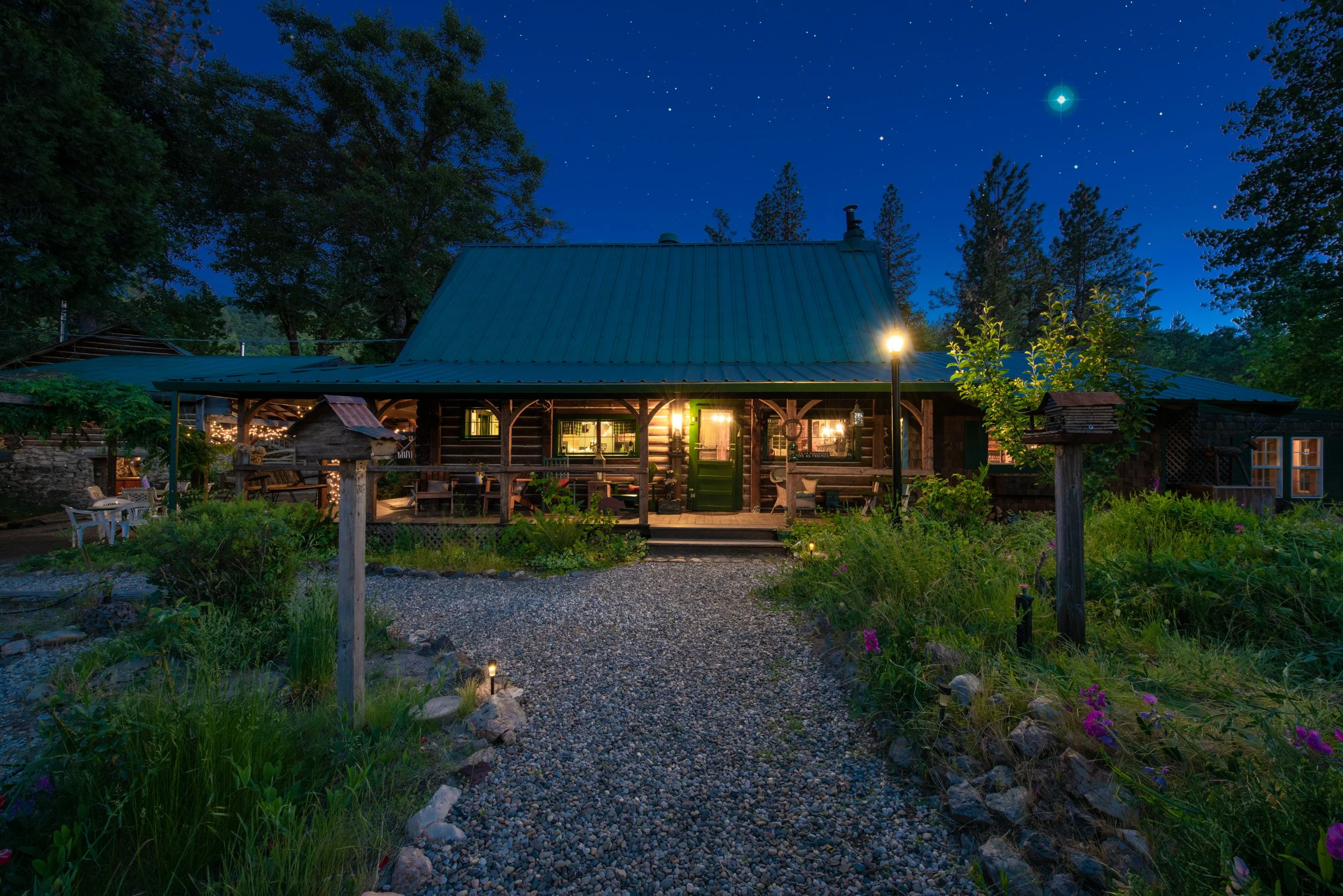 Nighttime photo of a cozy cabin with lights glowing inside, surrounded by trees and garden, under a dark starry sky with a bright planet or star visible.