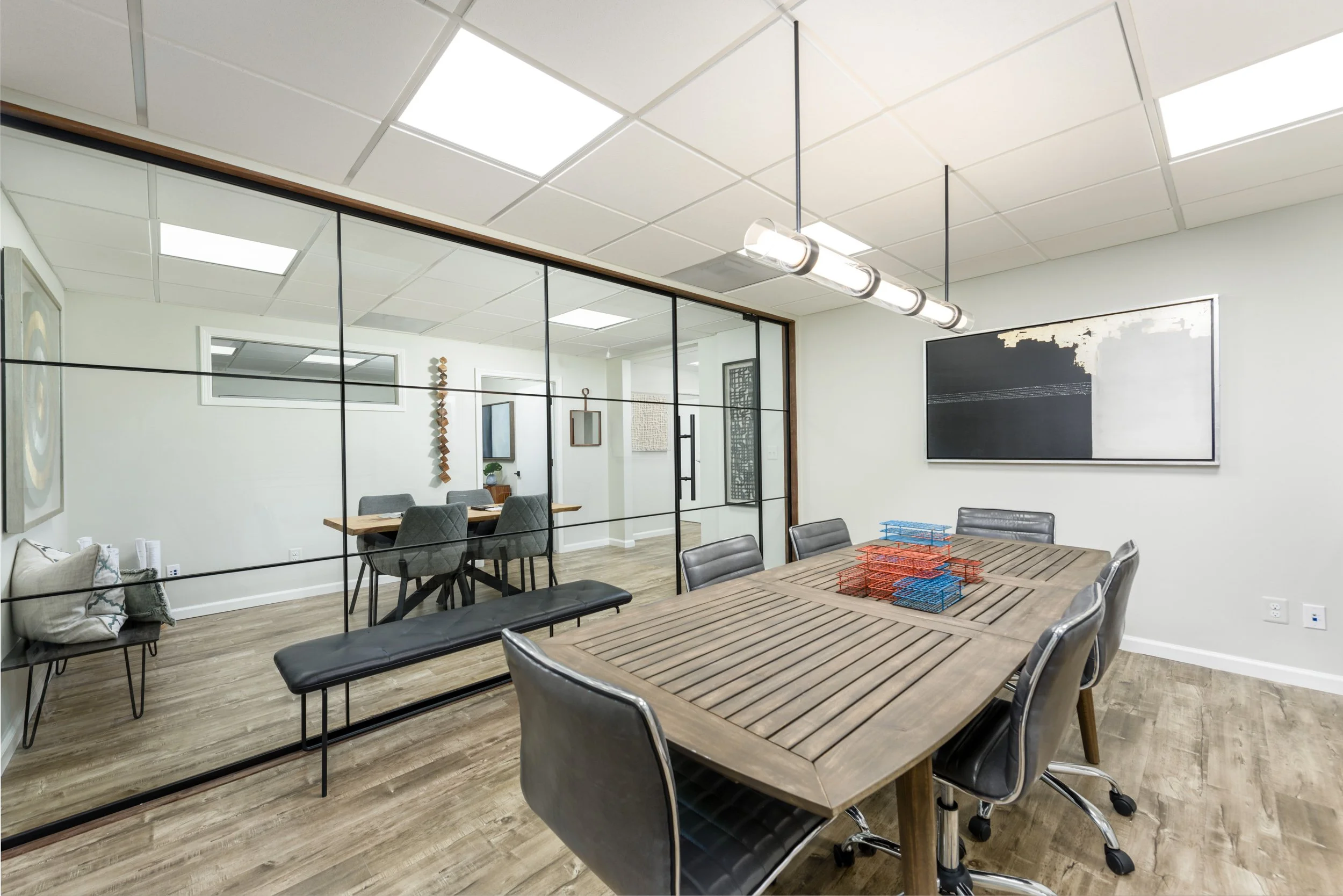 Conference room with a large wooden table, black chairs with metal bases, a pendant light, office artwork, and a glass wall separating it from another room.