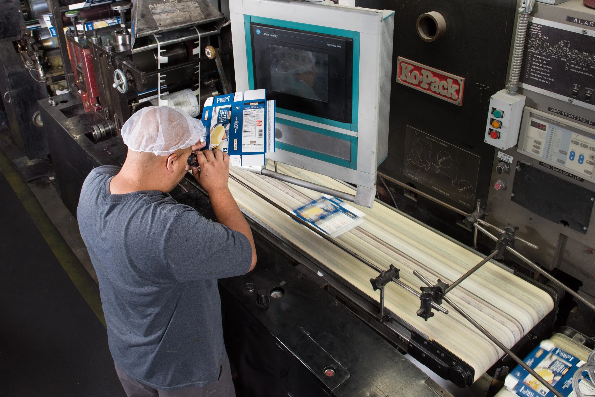 A worker in a hairnet operates machinery in a manufacturing facility, packaging boxes of butter onto a conveyor belt with a control panel nearby.