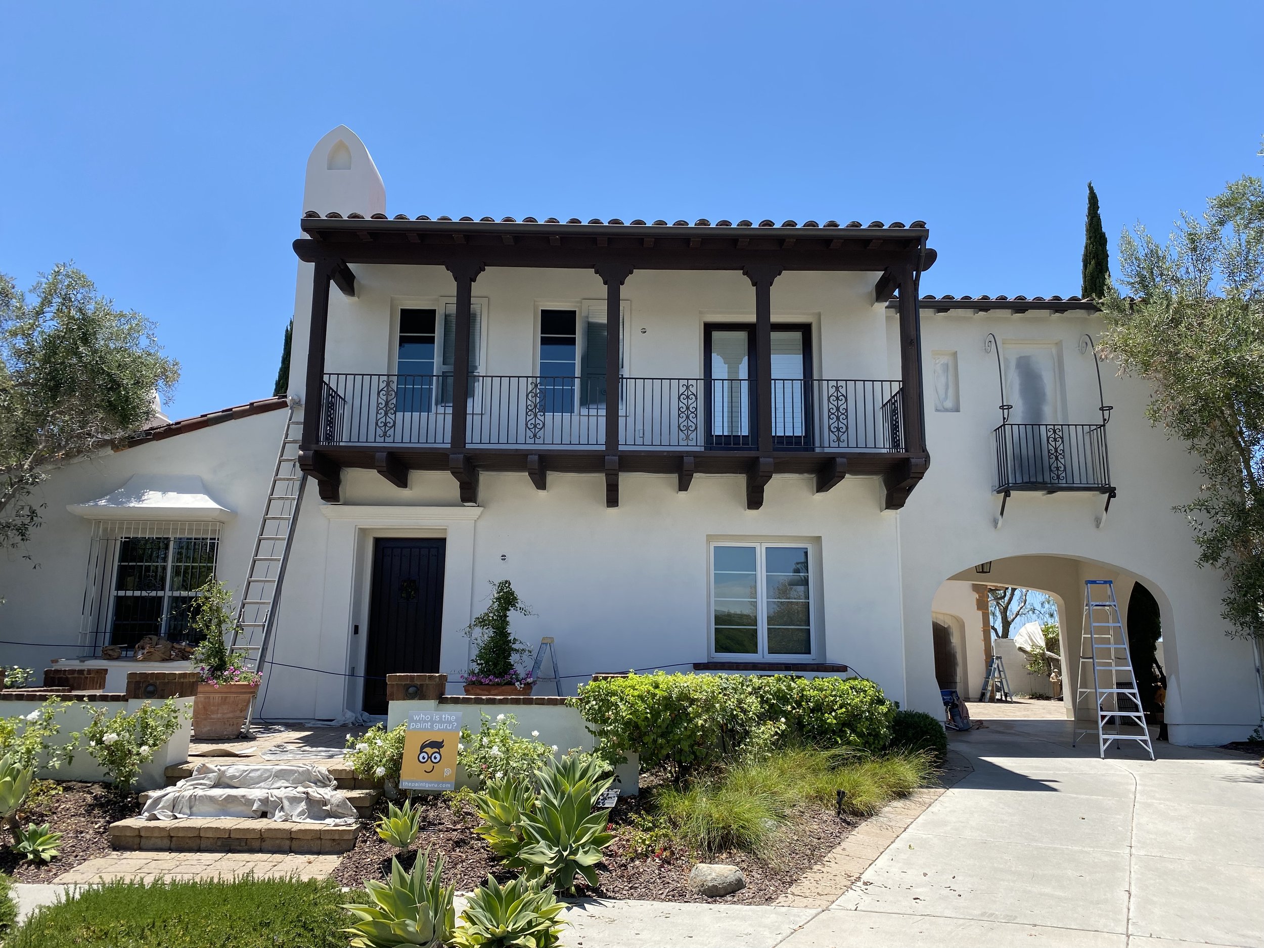 A two-story white house with a dark brown balcony on the second floor, surrounded by trees and plants, with ladders leaning against the house and construction signs indicating ongoing work.