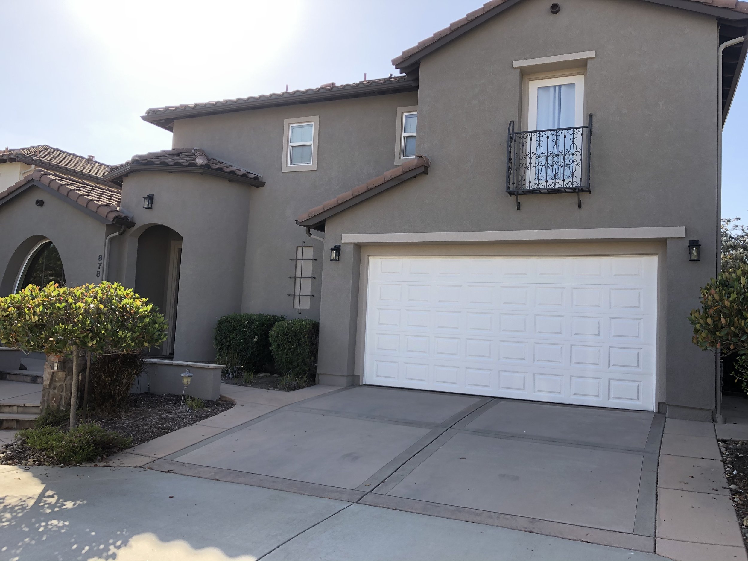 A two-story gray house with a white garage door, small balcony, tiled roof, and landscaped front yard.