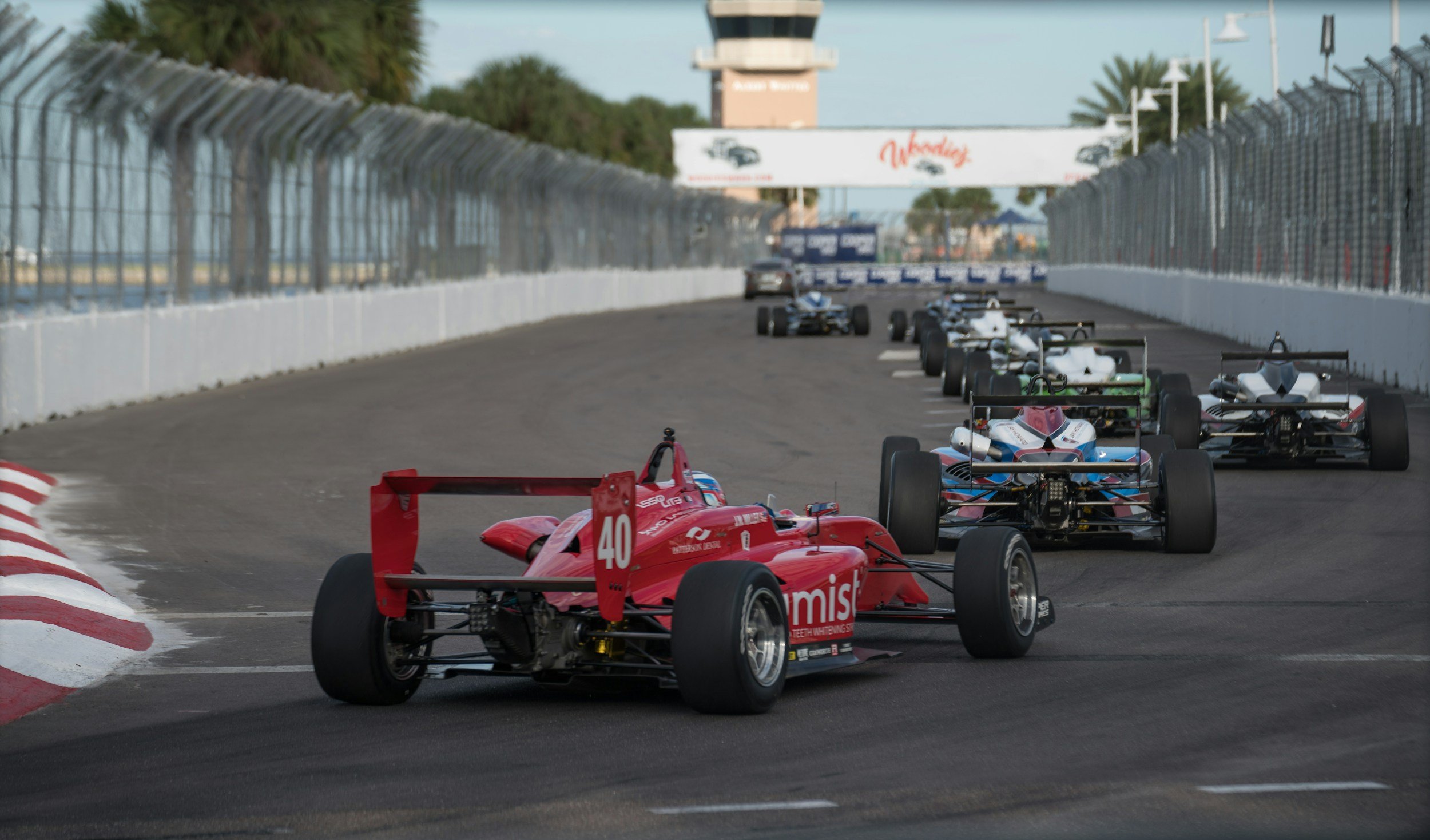 Race cars on a racing track, with a red car numbered 40 leading, followed by several black and white cars, and a blue car further back. The track is bordered by a white barrier with a fence on top, and a tall control tower can be seen in the background.