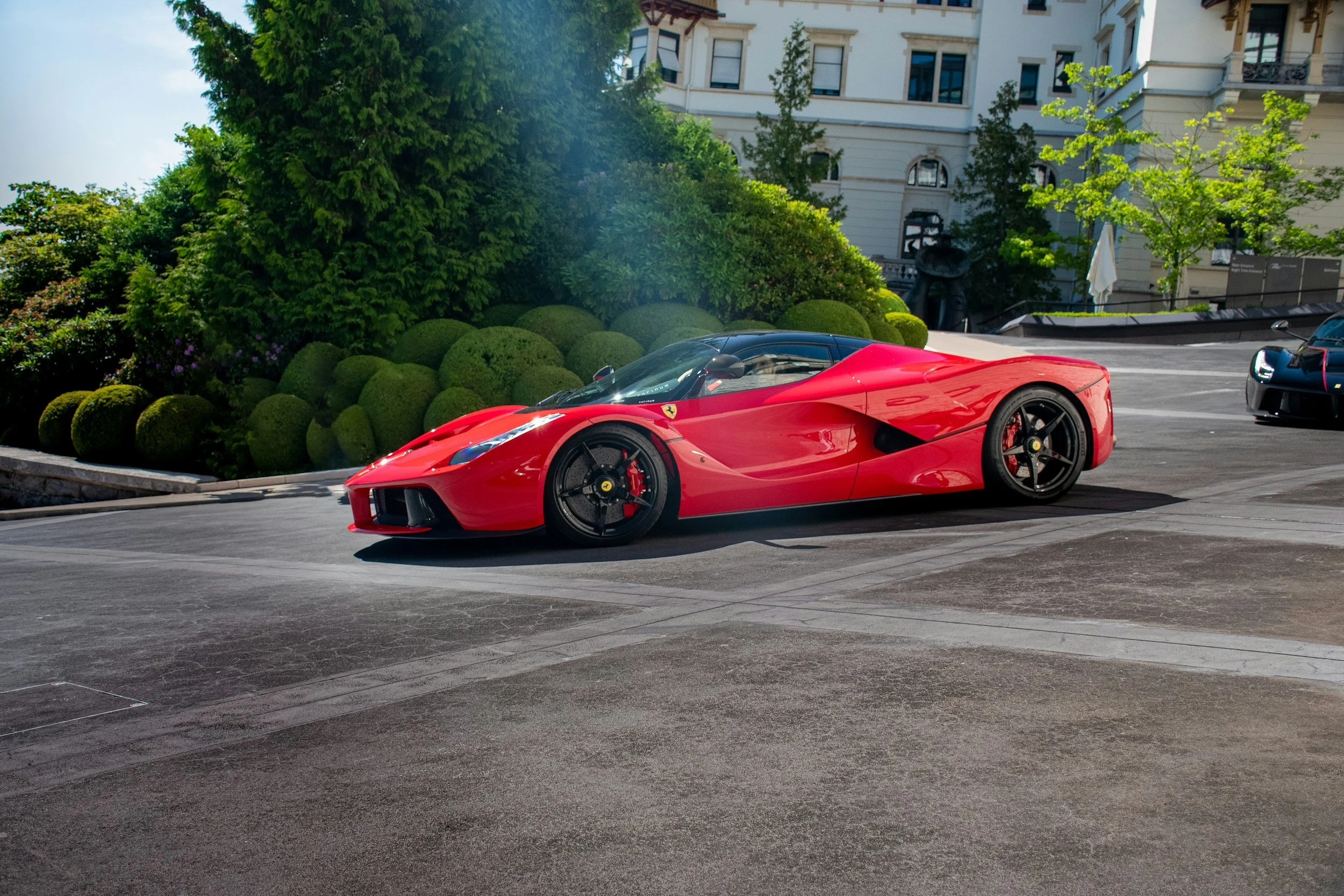 A red Ferrari sports car parked on a street with a building, trees, and bushes in the background.
