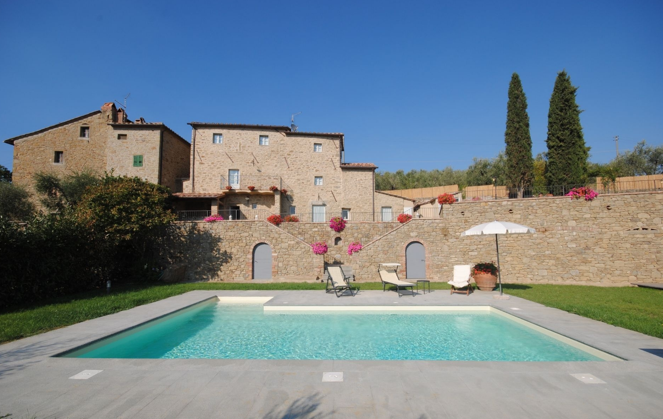 A swimming pool in front of a stone house with outdoor seating and colorful flowers under a clear blue sky.