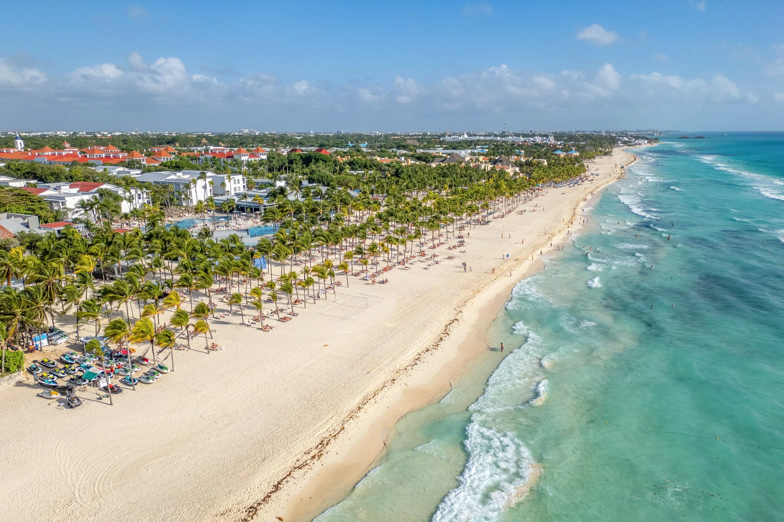 Aerial view of a beach with white sand, green palm trees, umbrellas, and sunbathers along the shoreline. There are ocean waves and clear turquoise water on the right, with a resort and buildings with red-tiled roofs on the left.