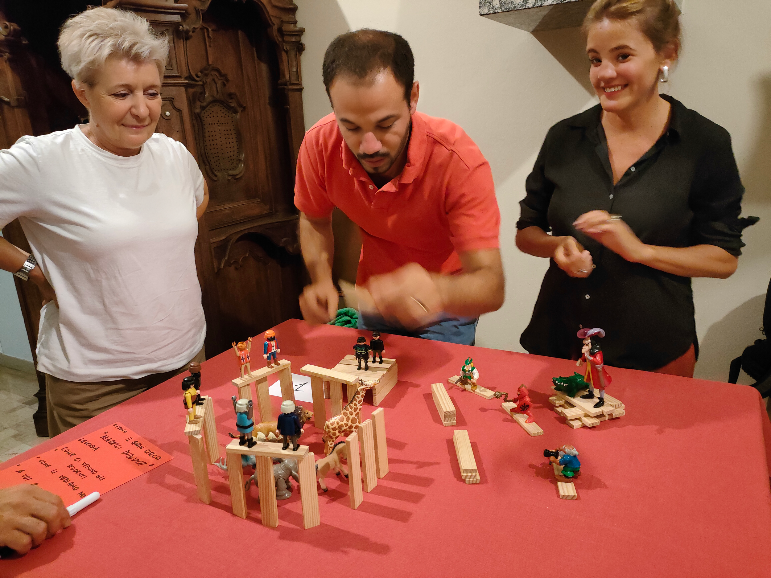 Tre persone osservano e interagiscono con un modello di costruzioni di legno su un tavolo rosso. Sono presenti figurine colorate e blocchi di legno disposti in modo creativo. Un cartello scritto a mano è visibile in basso a sinistra.