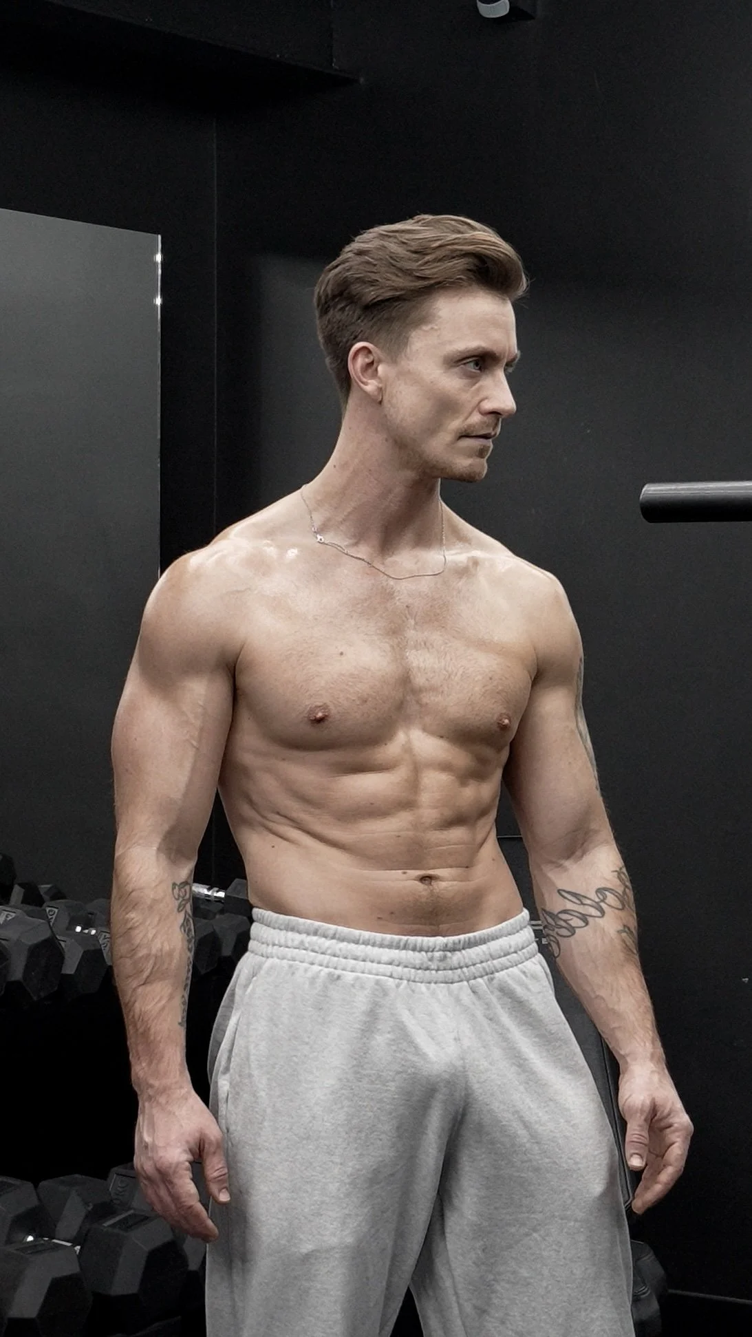 A shirtless man with a muscular physique and tattoos on his arms, wearing gray sweatpants, standing in a gym with black walls and dumbbells in the background.