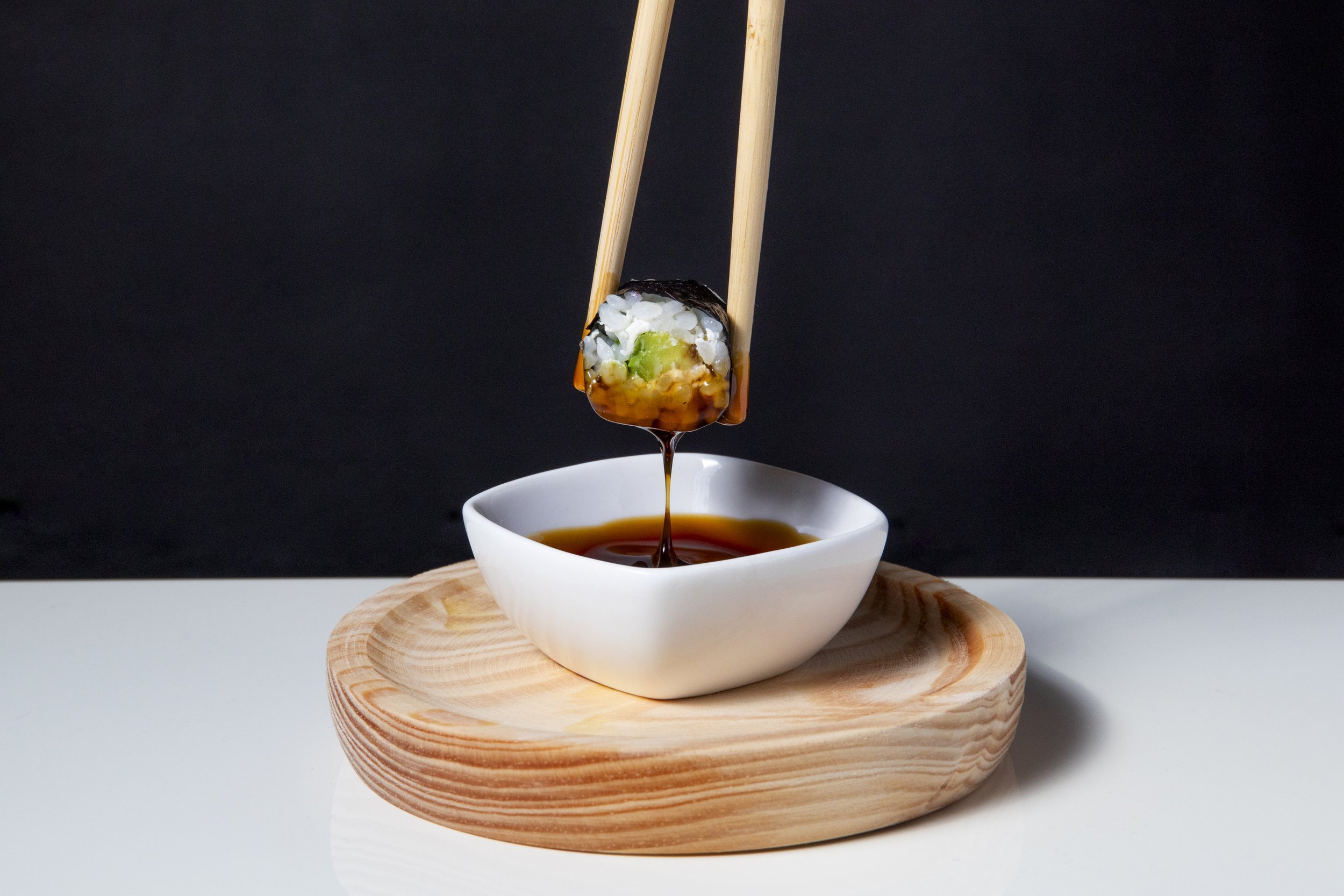 Un trozo de sushi con salsa de soja cayendo en un cuenco blanco sobre una base de madera.