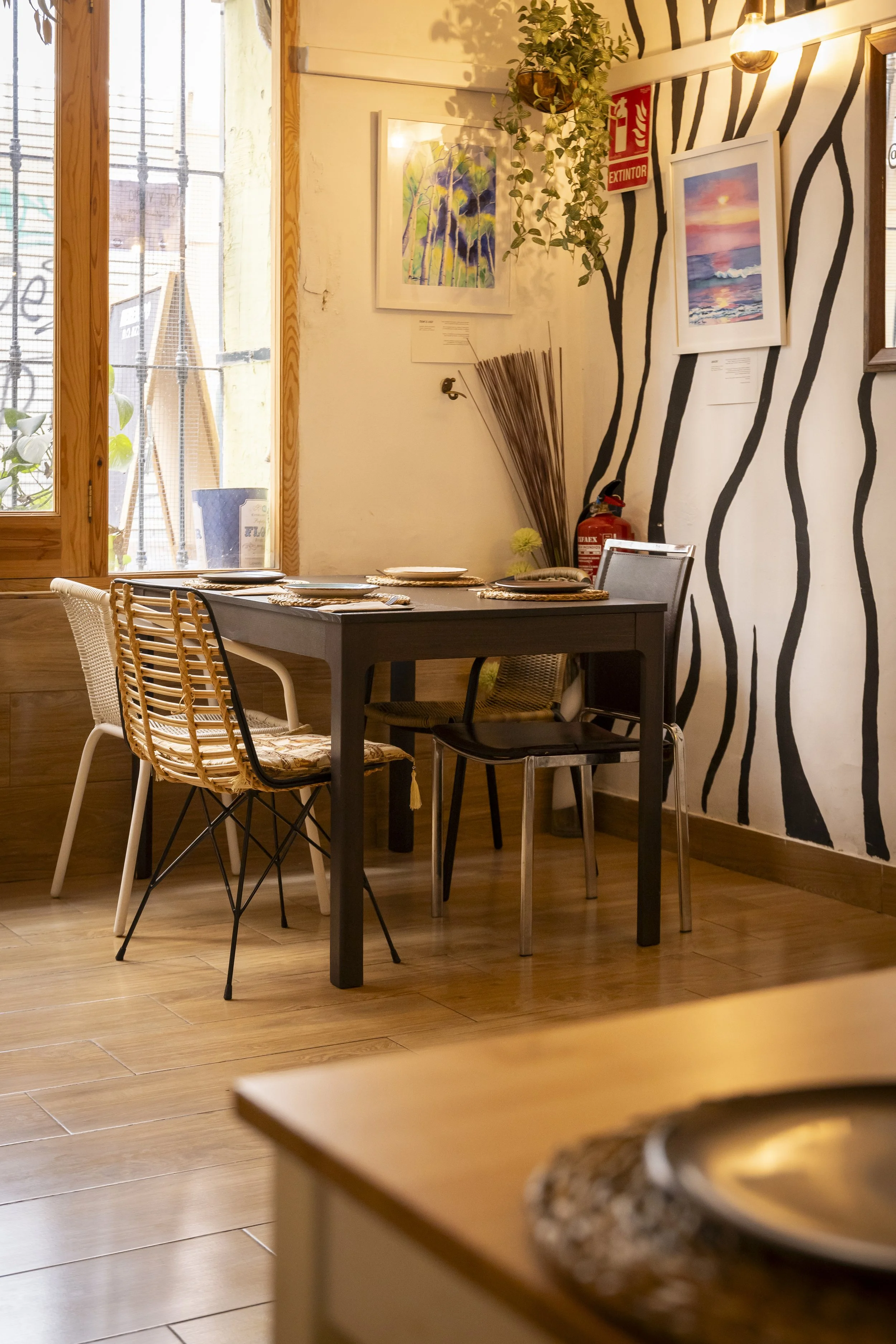 Interior de un restaurante con mesa de comer, sillas de varios estilos, paredes decoradas con pinturas y plantas, con luz natural entrando por la ventana.