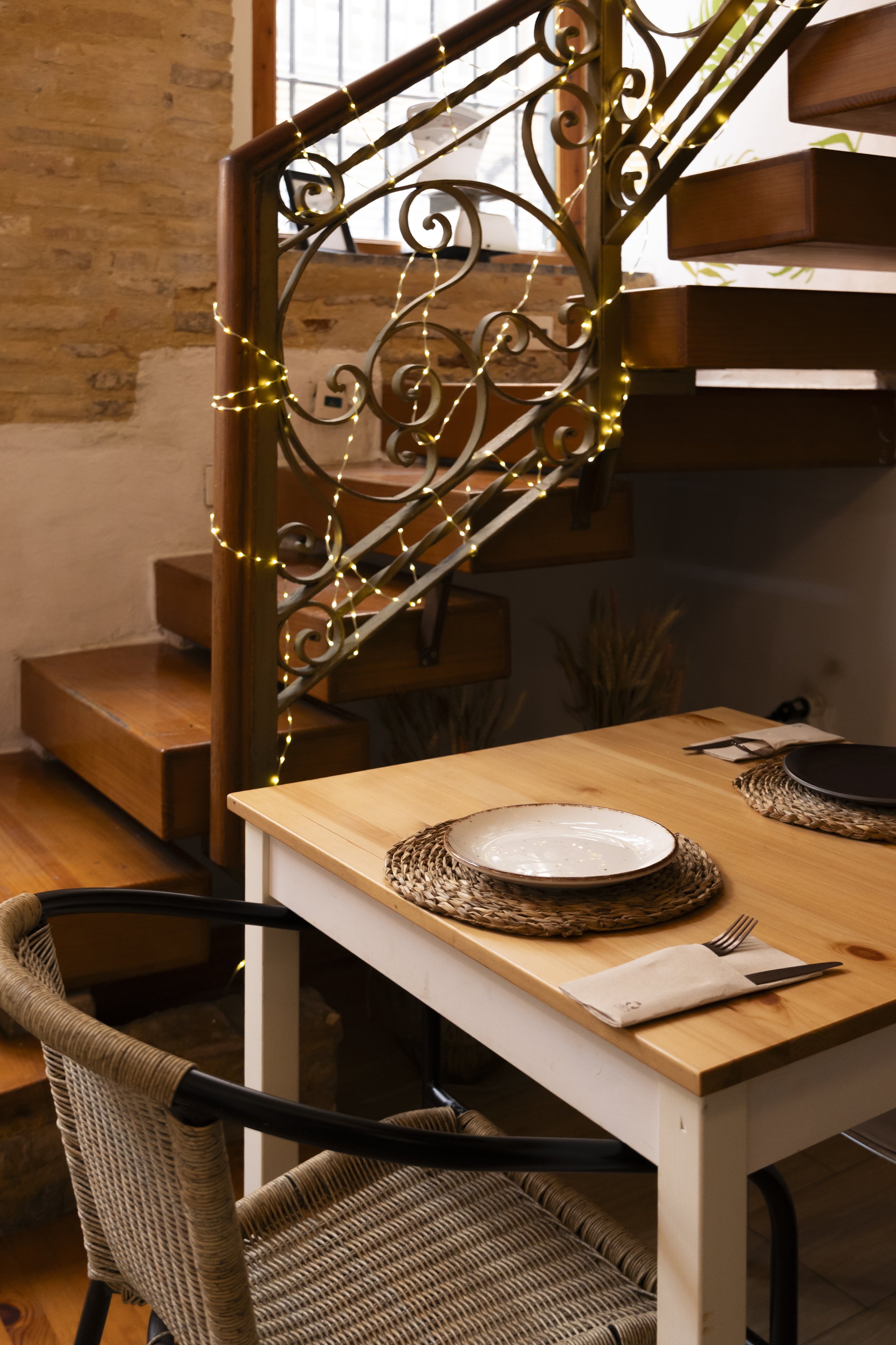 Mesa de comedor con platos, tenedores, cucharas y manteles de yute en un ambiente decorado con luces y una escalera de madera y hierro.