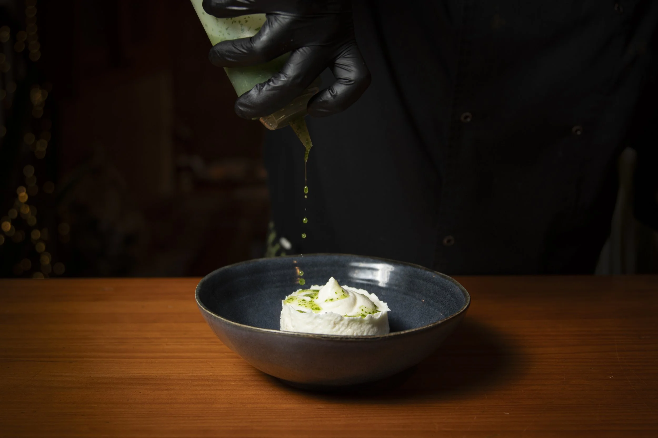 Una persona con guantes negros vierte líquido verde sobre un plato con comida blanco en un entorno de cocina o restaurante.