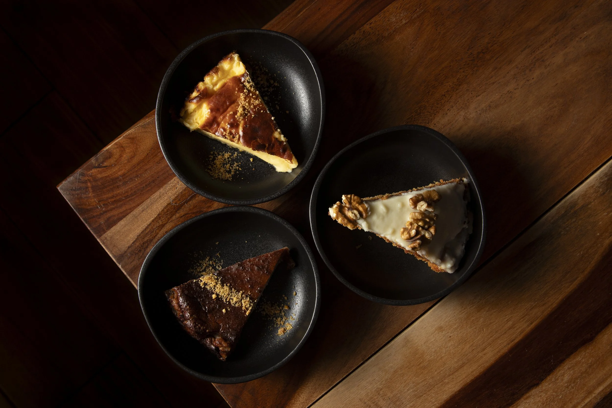 Tres rebanadas de diferentes pasteles en platos negros sobre mesa de madera.