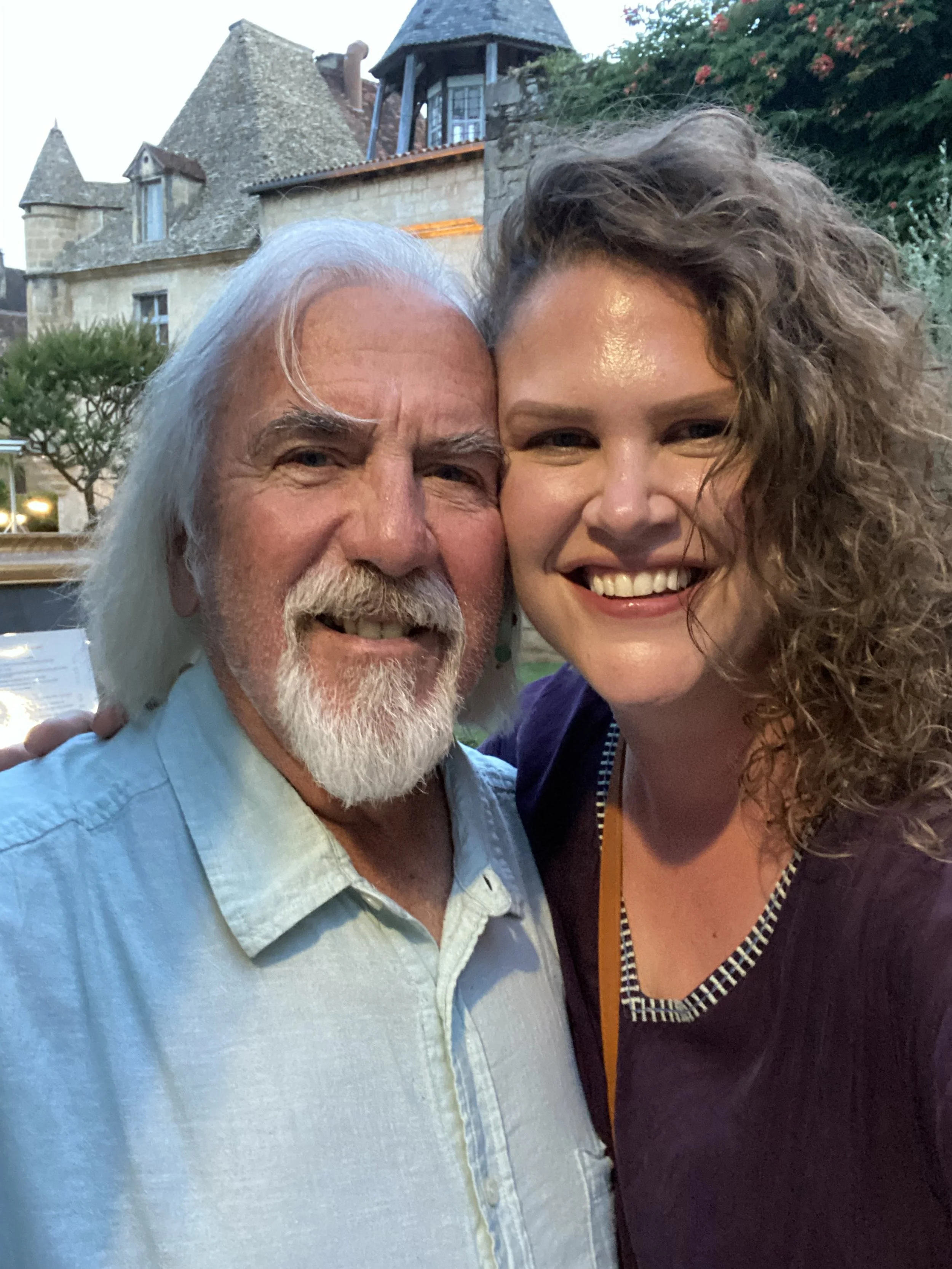 A man and a woman smiling and close together outdoors, with a historic stone building and greenery in the background.
