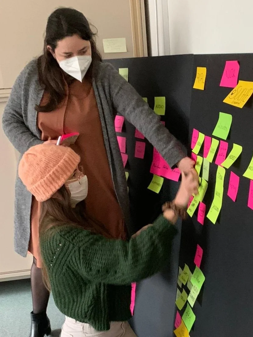 Zwei Frauen, eine ältere und eine jüngere, arbeiten mit bunten Post-its an einer Pinnwand. Beide tragen Masken, die ältere Frau hat dunkle, die jüngere trägt eine orange Mütze.