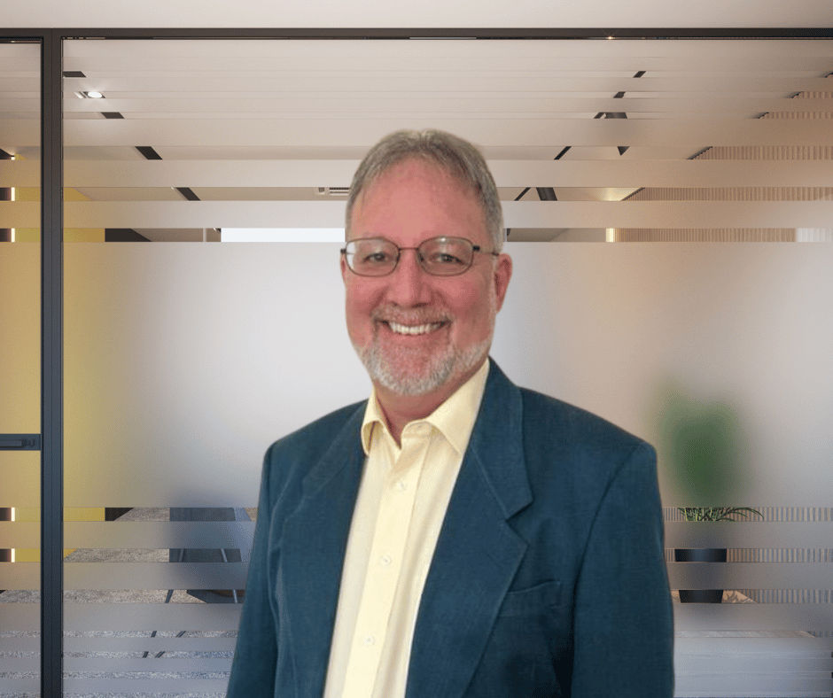 A smiling man with glasses, a beard, and short gray hair, dressed in a yellow shirt and a blue blazer, standing in an office with glass walls.