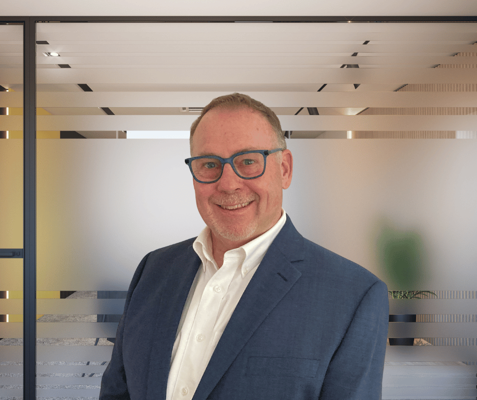 A man with glasses and a beard smiling, wearing a dark blue suit and white shirt, standing in an office with frosted glass walls and modern ceiling design.