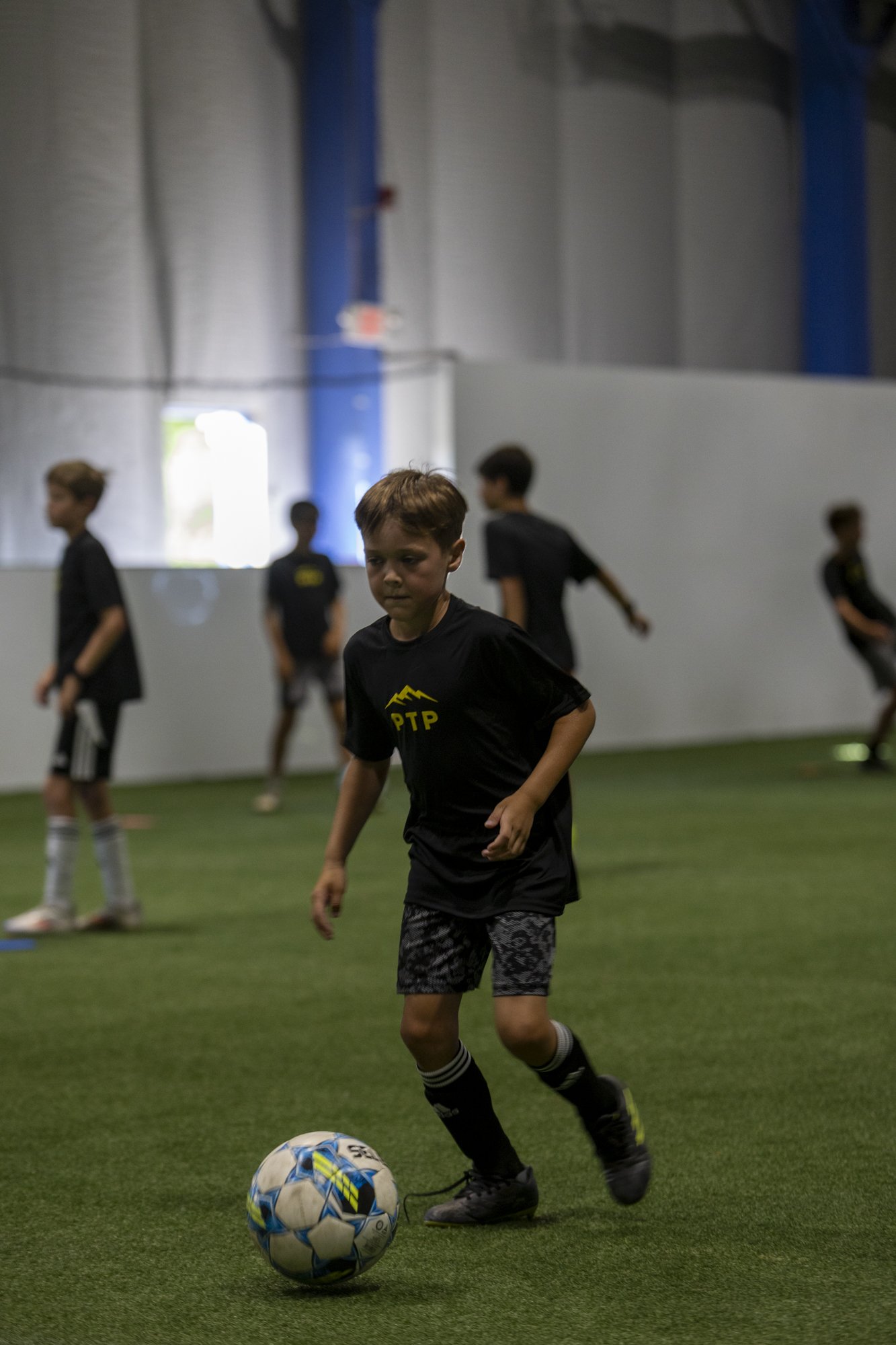 A young boy playing soccer indoors on a green turf field, surrounded by other boys in black jerseys.