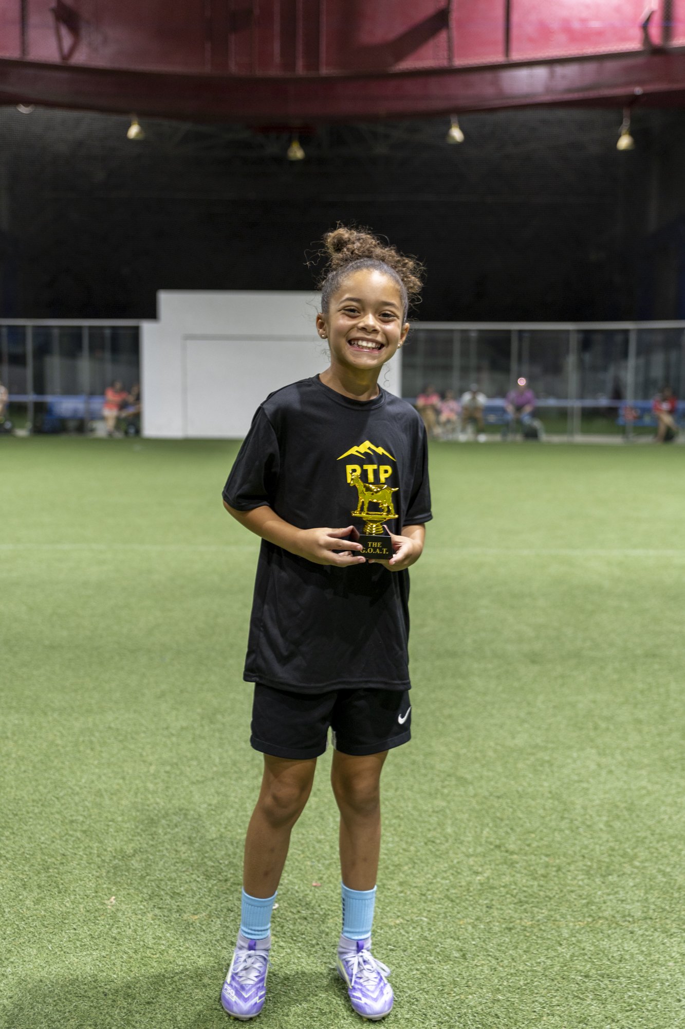 Young girl holding a trophy on a sports field, smiling, wearing a black sports uniform and purple cleats.