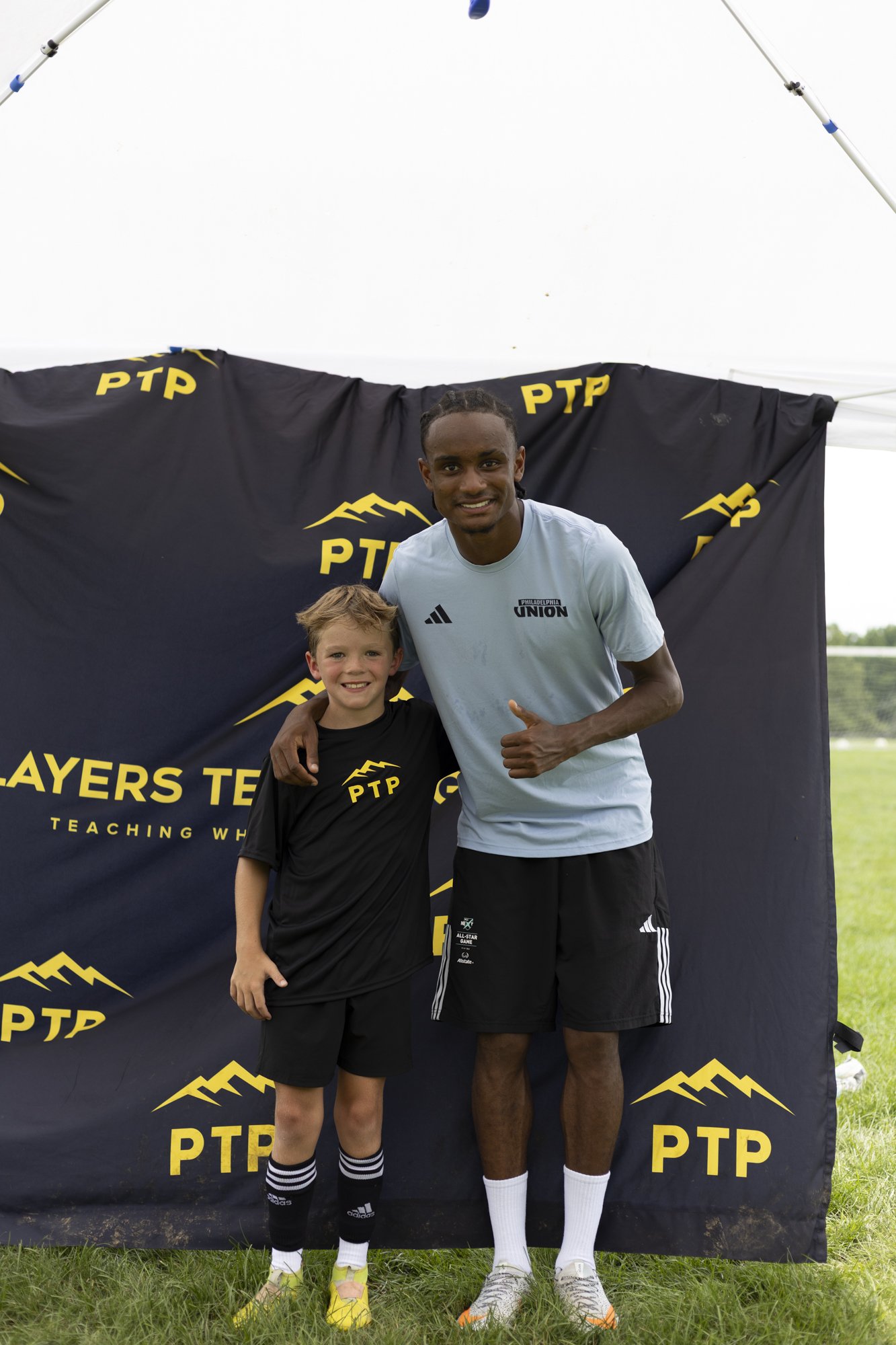 A man and a young boy, dressed in athletic clothing, standing together outdoors in front of a black backdrop with yellow logos for PTP and Philadelphia Union, both smiling at the camera.