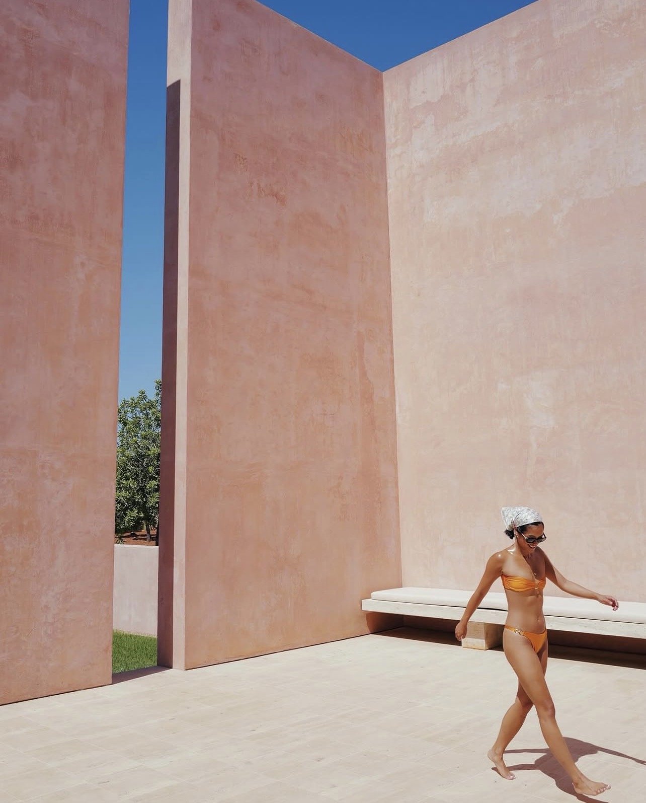 A woman in an orange bikini, sunglasses, and a white headscarf walking on a beige tiled surface next to a modern building with pinkish-orange walls and a clear blue sky.