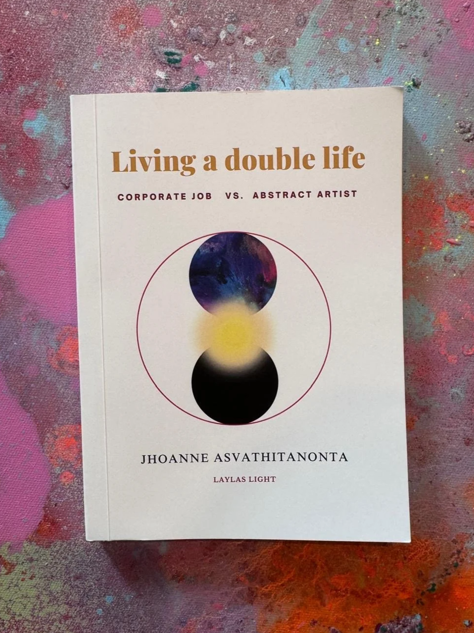Book titled 'Living a double life: Corporate job vs. abstract artist' by Jhoanne Asvathitanonta, on a colorful, painted surface.