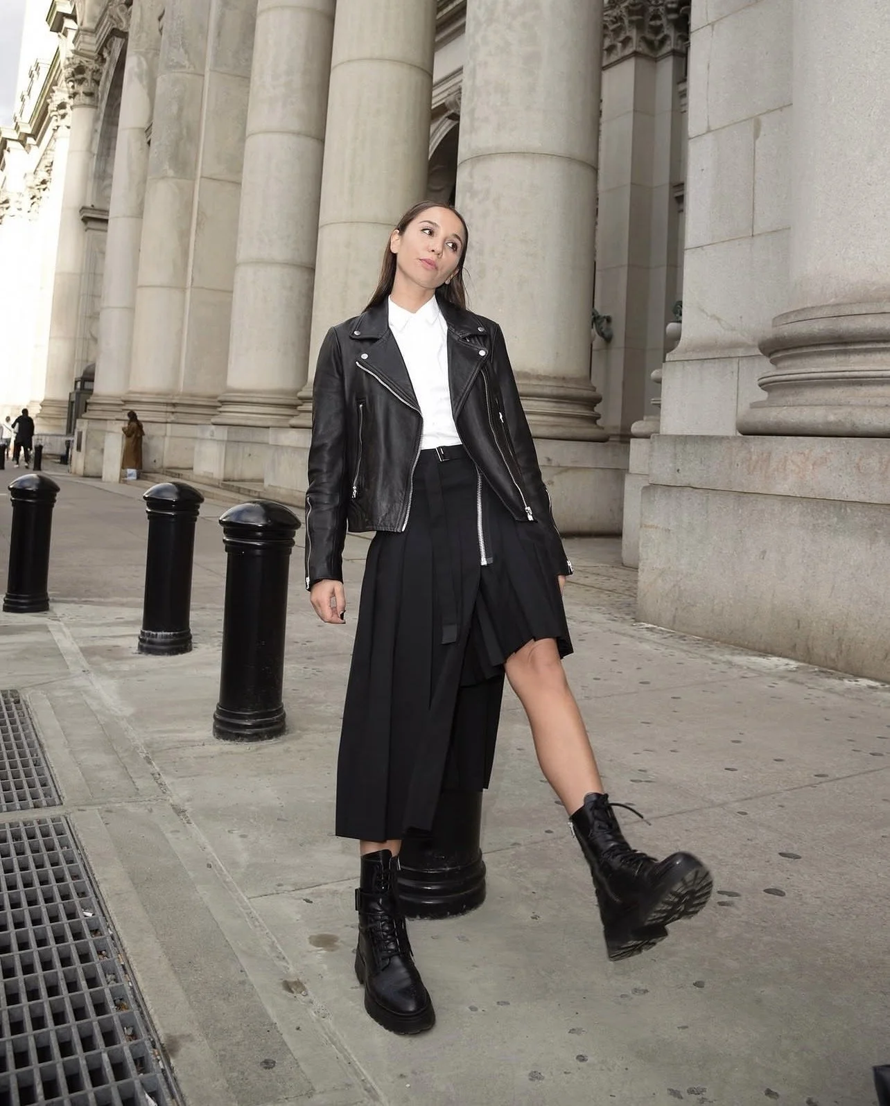 A young woman standing on a city sidewalk in front of large stone columns. She is wearing a black leather jacket, a white shirt, a long black skirt, and black combat boots. One leg is resting on a black bollard, and she has a confident expression.