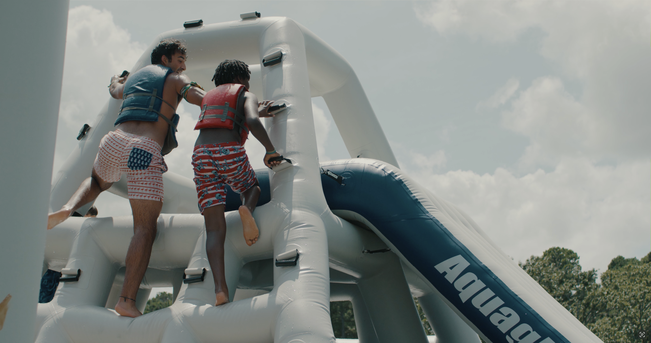 Two people wearing life jackets climbing an inflatable water slide labeled 'Aquaglide' on a sunny day.