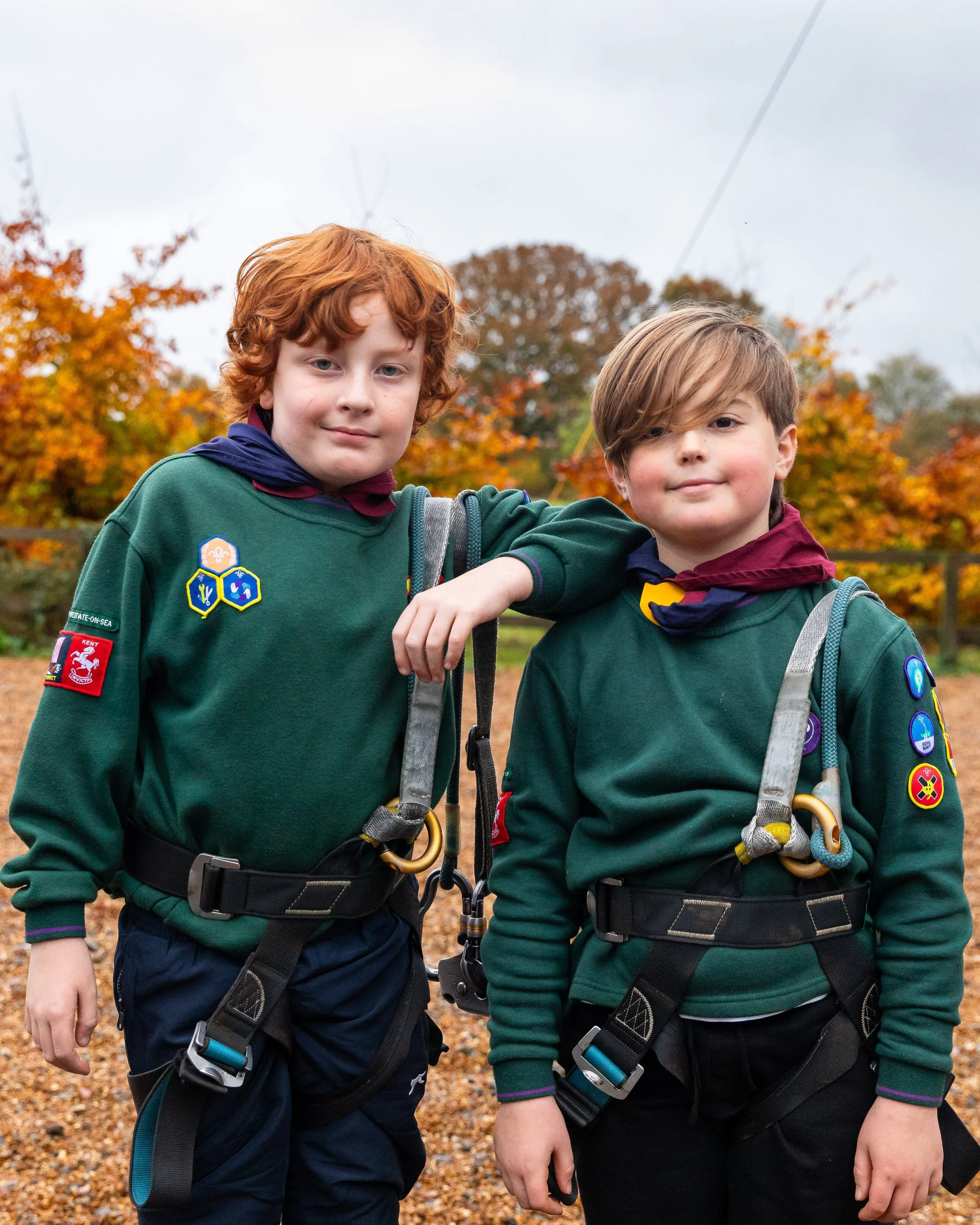 Two young boys wearing Scout uniforms with patches, standing outdoors during autumn with fall foliage in the background.