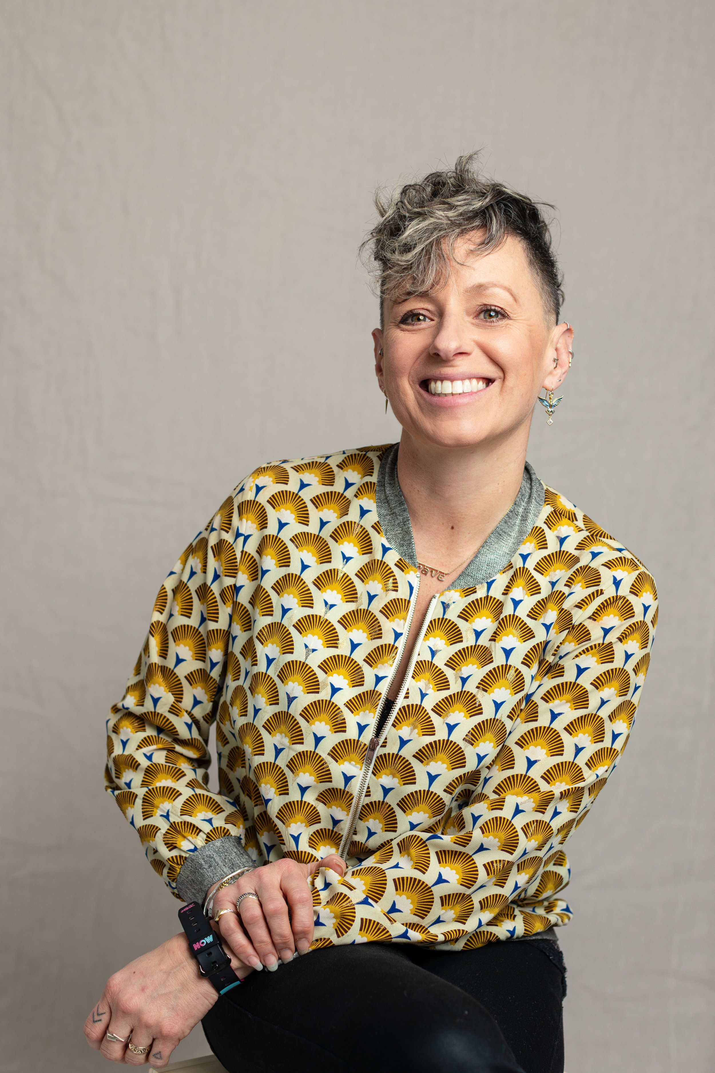 A woman with short, gray and black hair, smiling, wearing a patterned bomber jacket and sitting against a plain gray background.