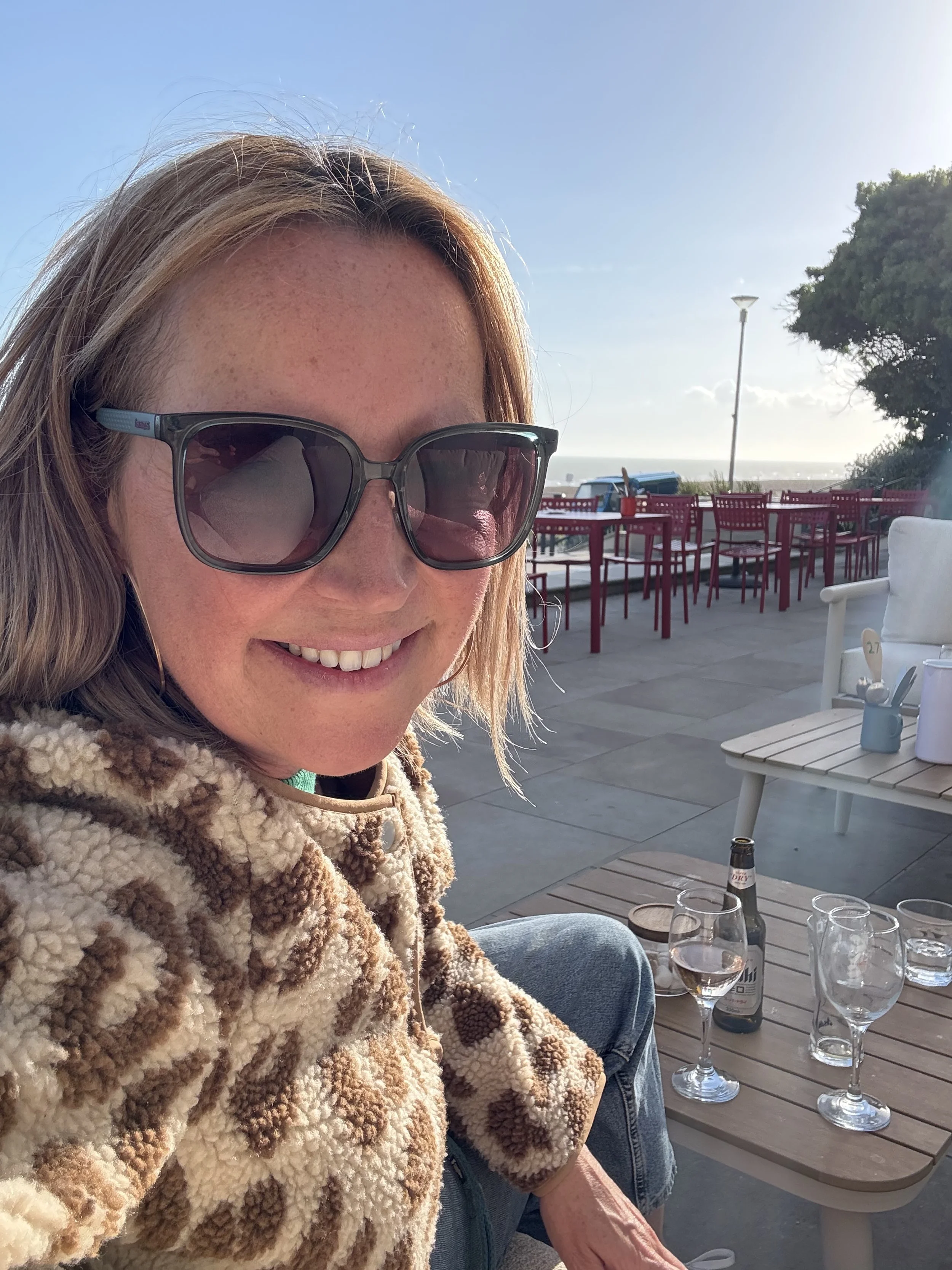 A woman with sunglasses sitting outdoors at a table with empty glasses and a beer bottle, smiling at the camera.