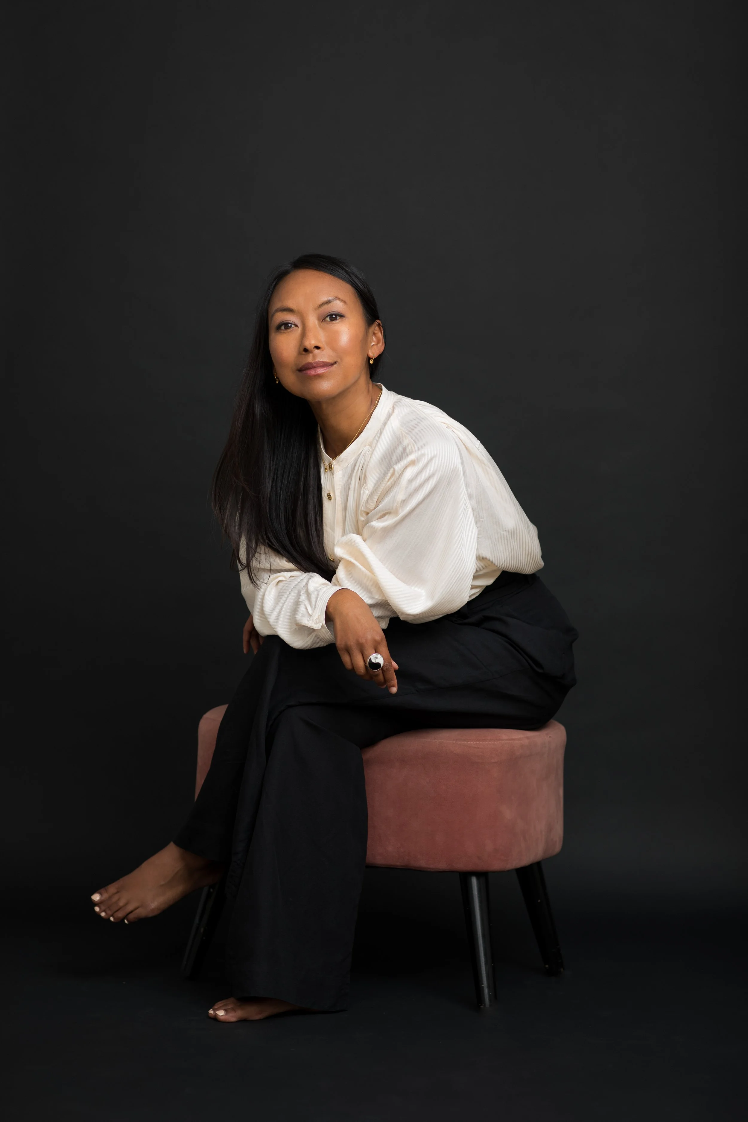 A woman with long black hair, wearing a white blouse and black pants, sitting on a pink cushioned stool against a black background.